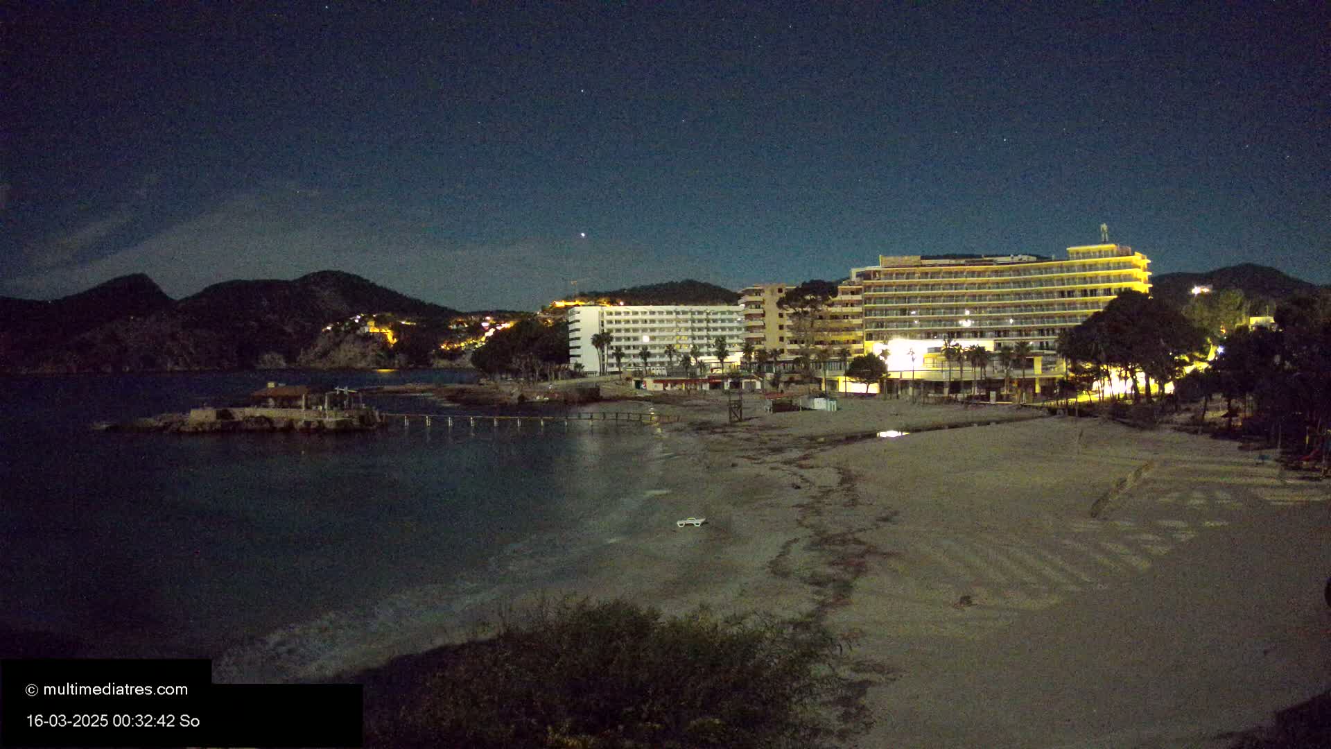 A nighttime view of a beach with hotels and a small island under a clear, starry sky.