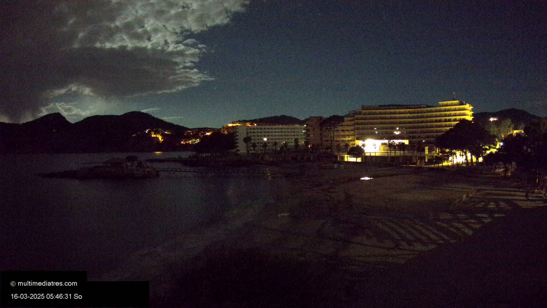 A nighttime beach scene shows a dark body of water, a dark sandy shore, and illuminated hotels and hills under a partly cloudy sky.