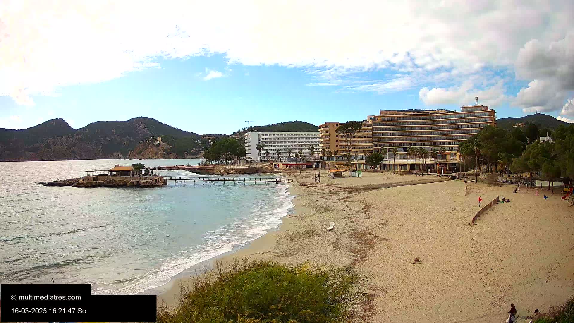 A sandy beach with calm, light blue water laps at its edge, featuring a small jetty, several hotels, and mountains in the background under a partly cloudy sky.