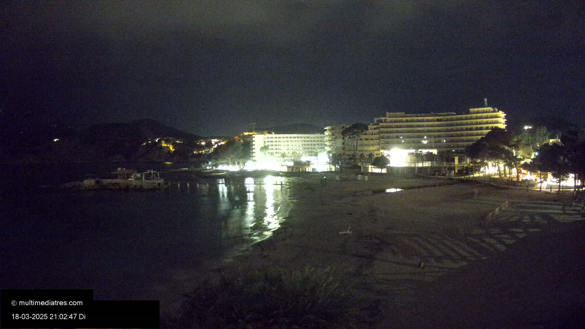At night, a dark beach with a calm body of water reflects the lights of several illuminated hotels and buildings on the shore, under a dark sky.