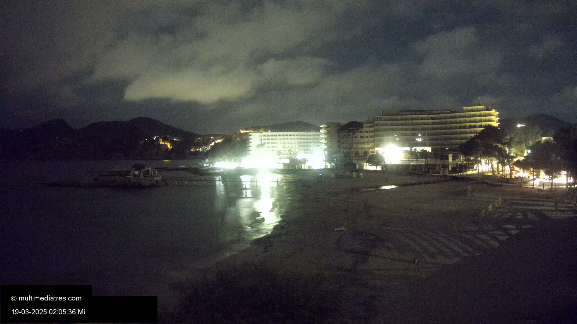 At night, under a cloudy sky, illuminated hotels are situated near a dark beach and calm body of water, with mountains in the background.