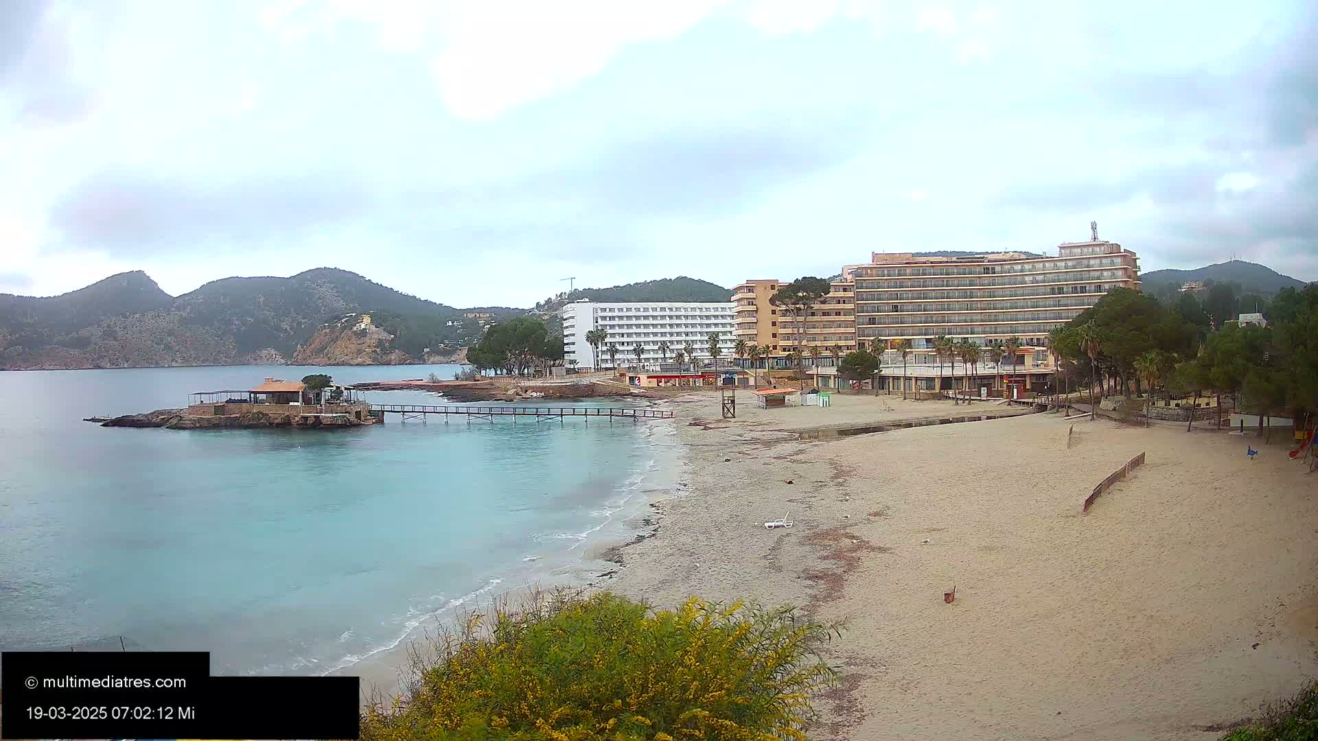 A sandy beach with calm, light blue water stretches towards two large hotels under a cloudy sky, backed by hills.