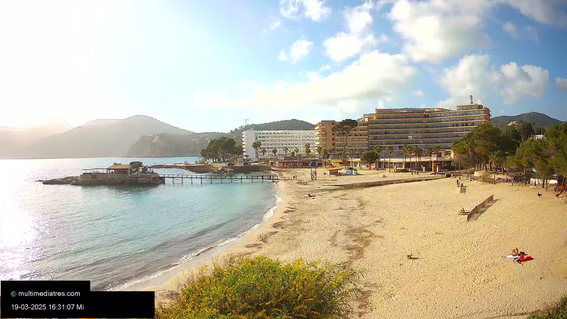 A sandy beach with several buildings and a pier extends into a calm, blue bay, backed by mountains under a partly sunny sky.