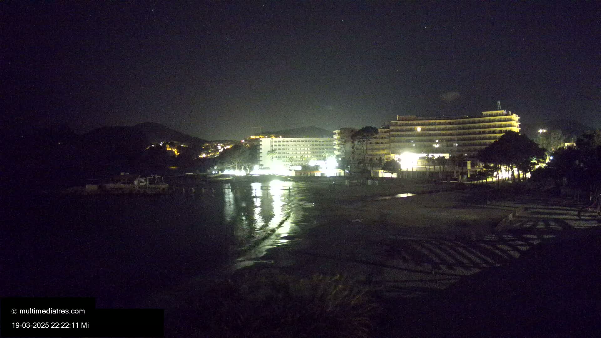 At night, two brightly lit hotels stand on a dark beach next to a calm, reflective body of water, with a hill visible in the background under a starry sky.