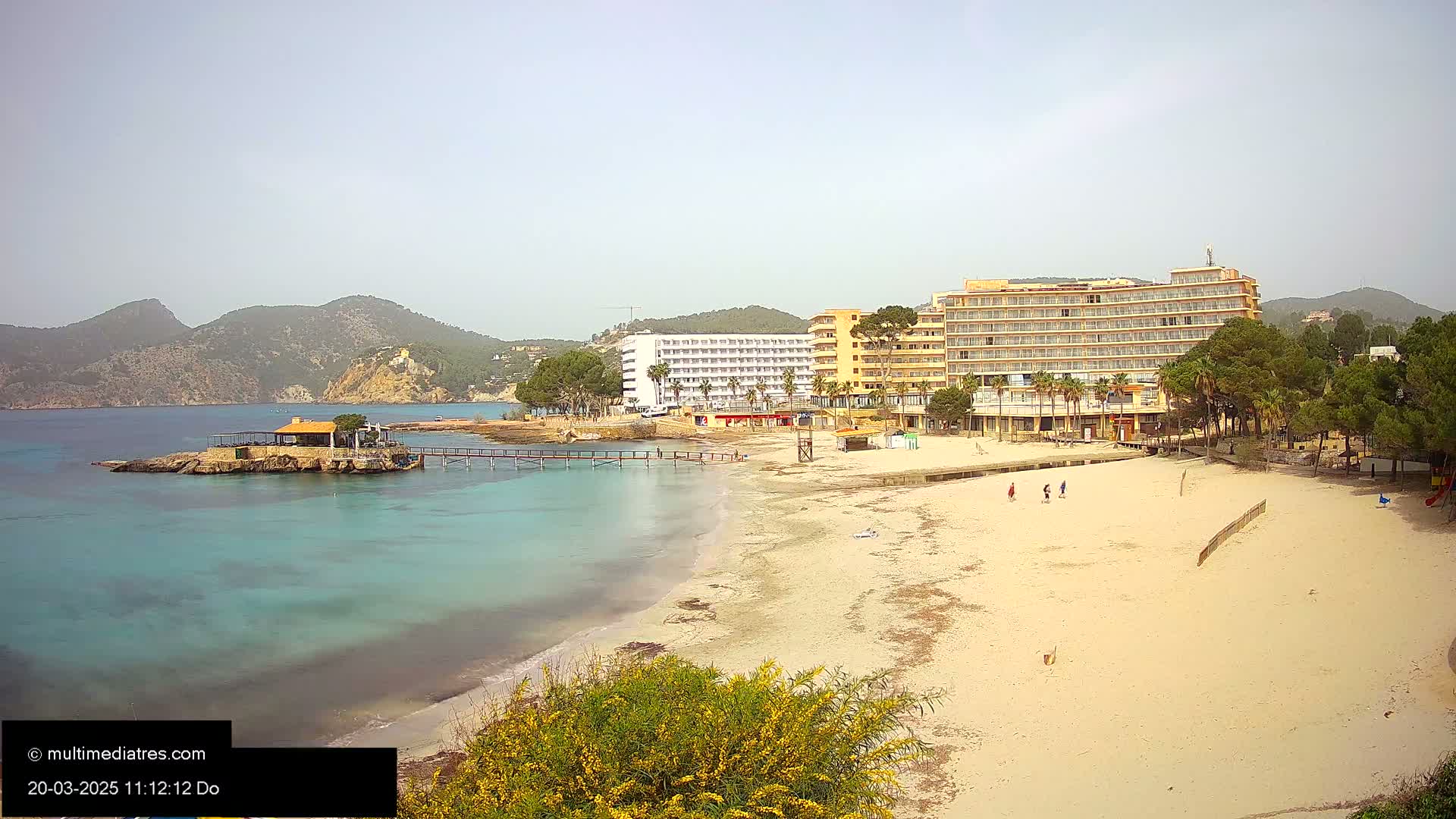A sandy beach with turquoise water, a pier, several hotels, and low-lying mountains under a hazy sky.