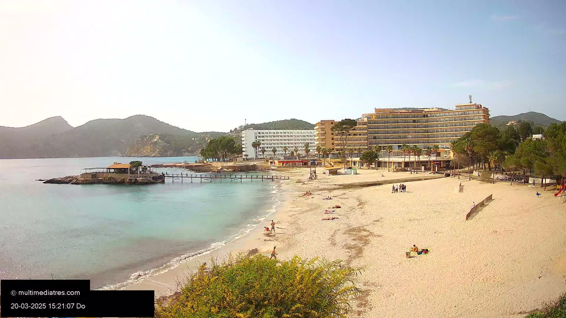 A sandy beach with a few people on it stretches out in front of several hotels and palm trees, under a clear blue sky.