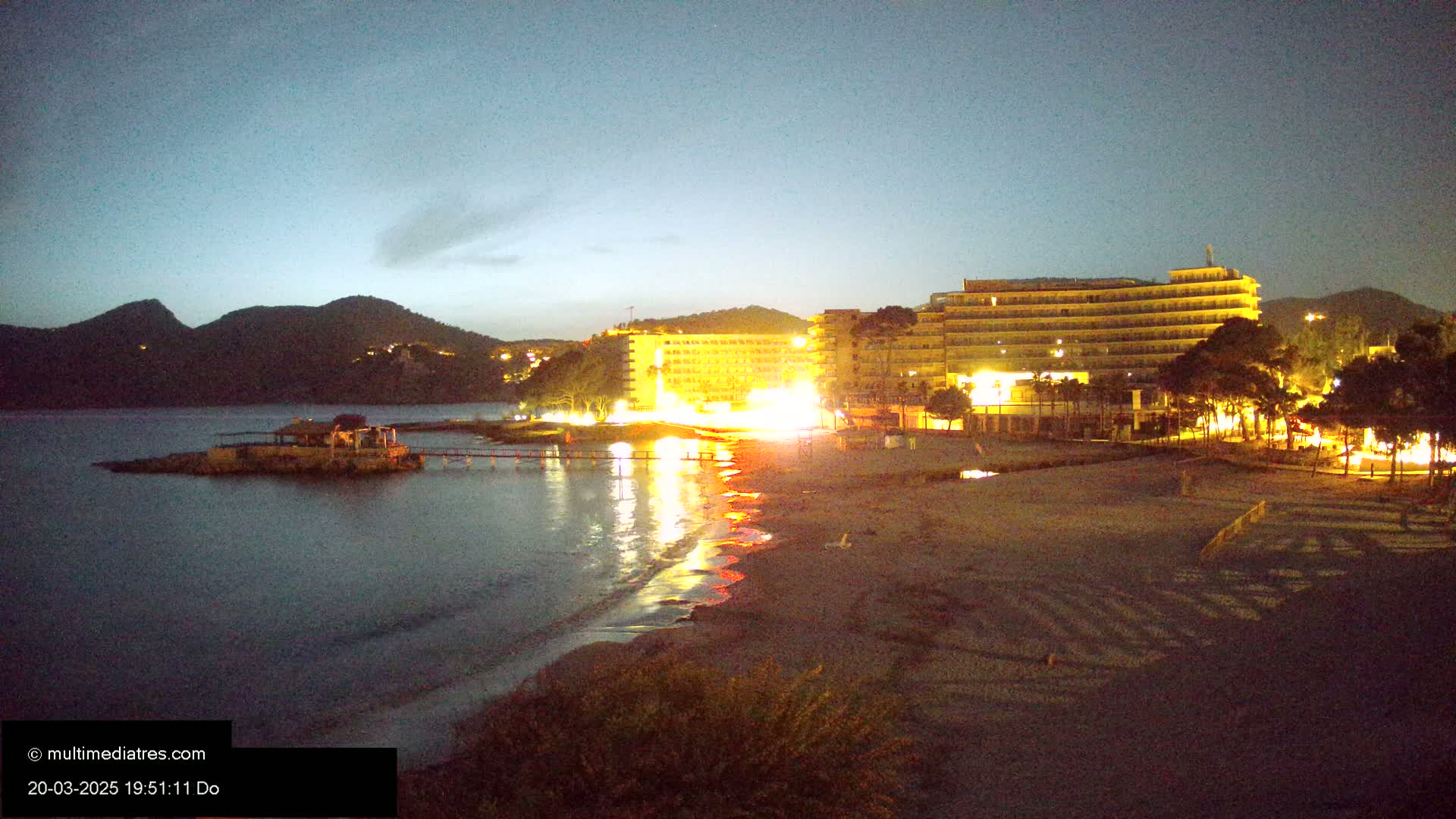 A sandy beach at night is illuminated by the lights of hotels and buildings behind it, with calm water in front and dark mountains in the background under a dusky sky.