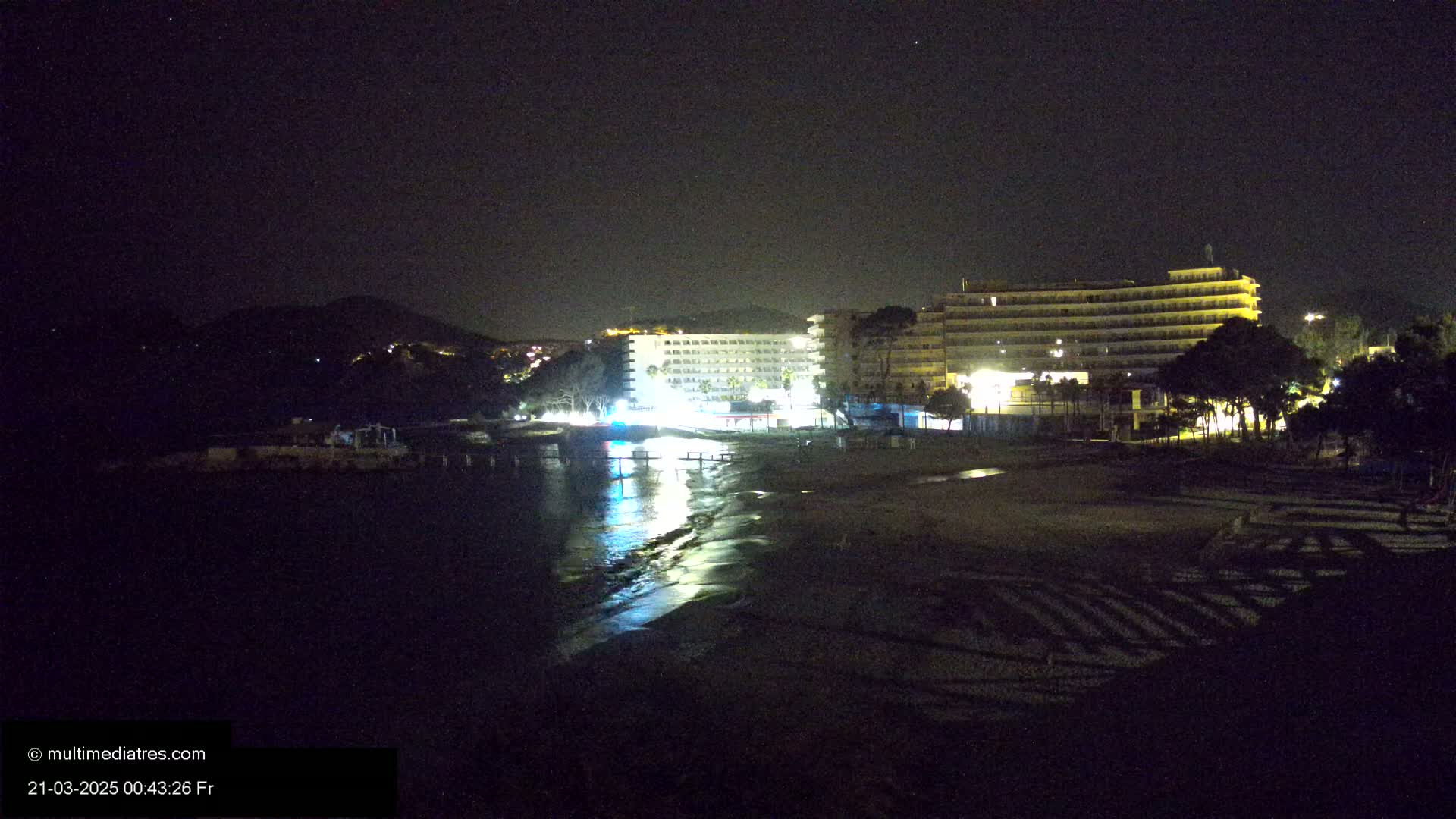 At night, two brightly lit hotels stand near a dark beach with calm water reflecting their lights, and a hill is visible in the distance under a clear, starry sky.