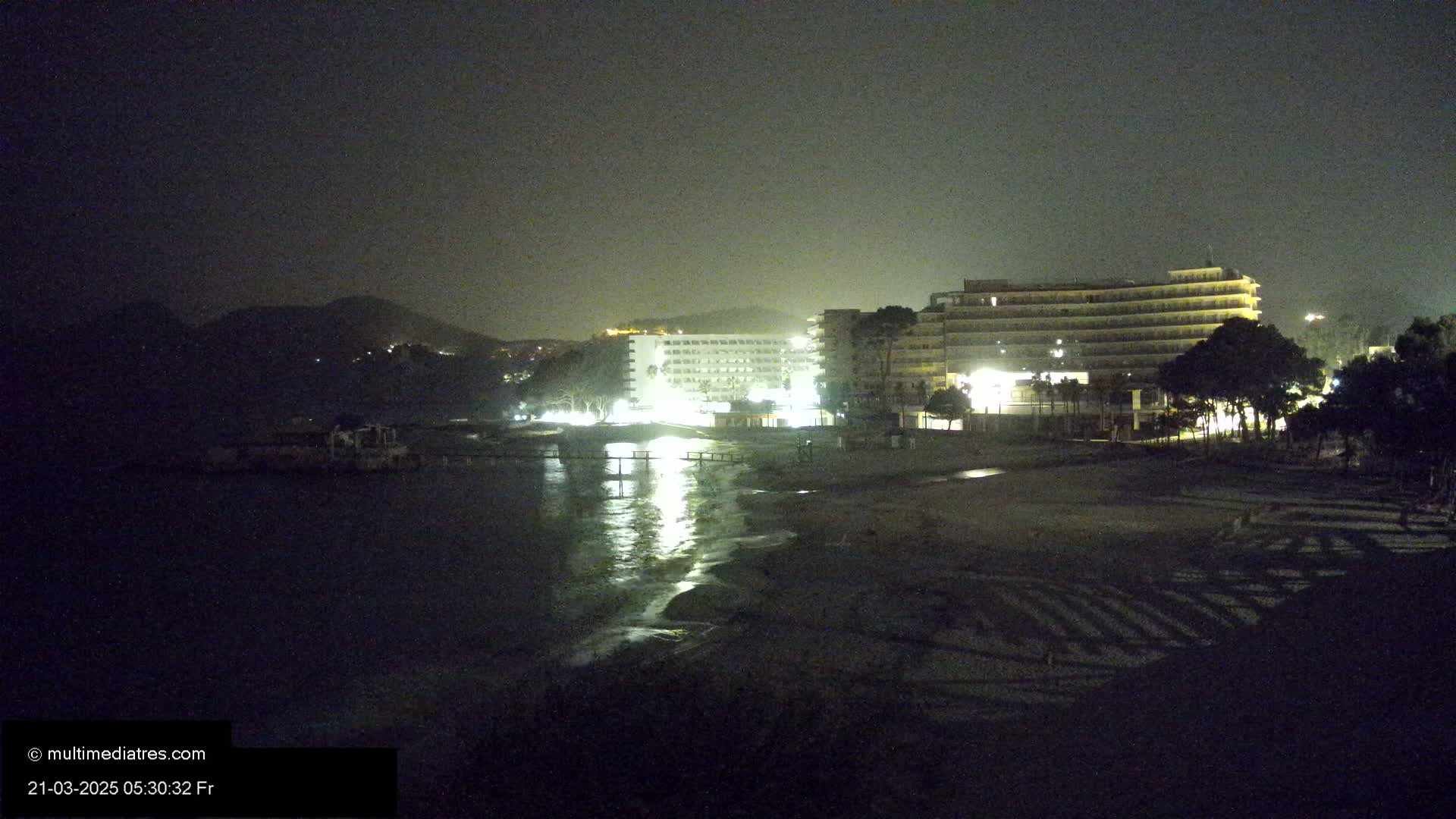 At night, two brightly lit hotels stand on a beach next to a calm, dark sea under a dark sky.