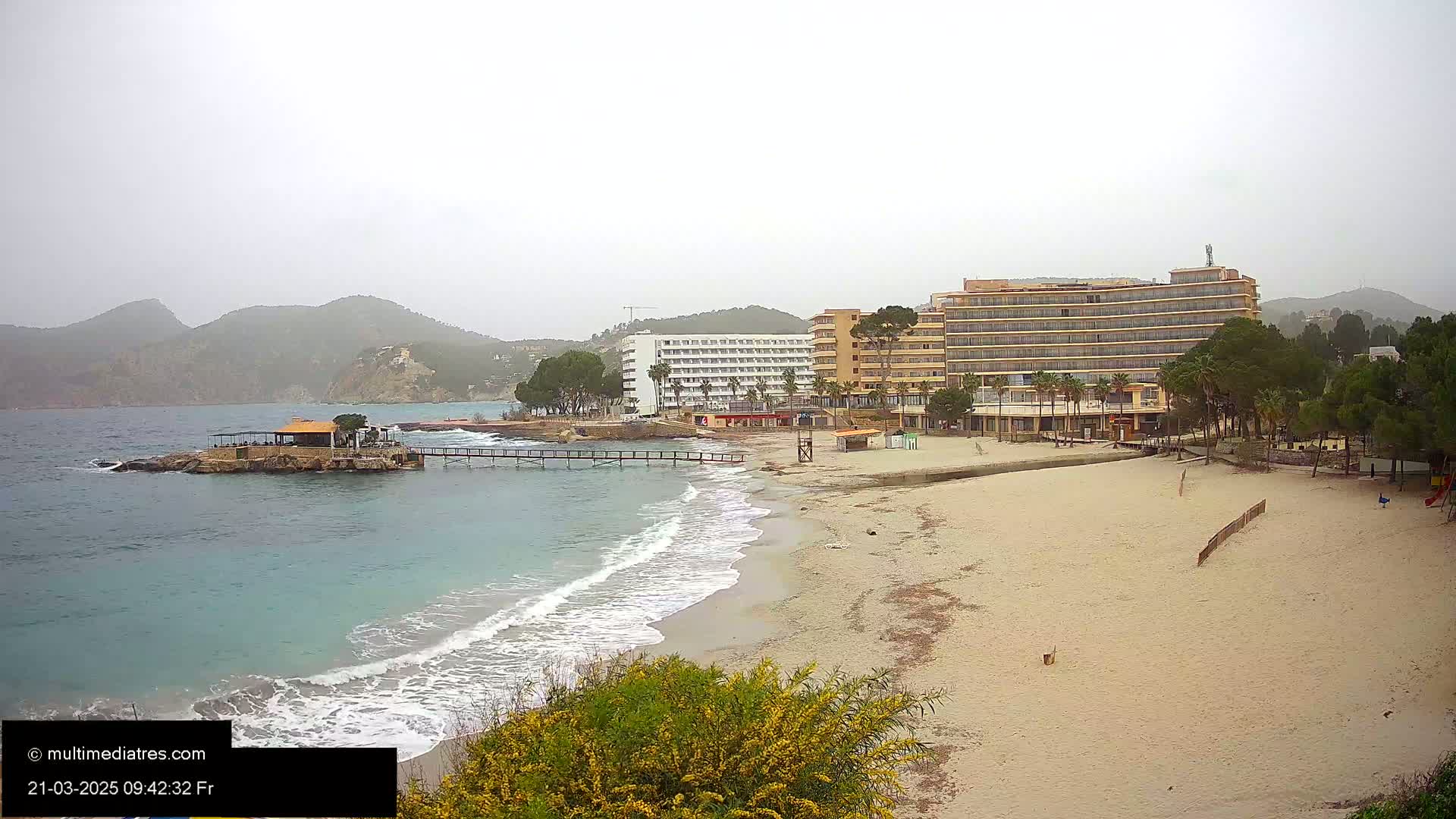 A sandy beach with a calm sea, hotels, and a small island with a building under an overcast sky.
