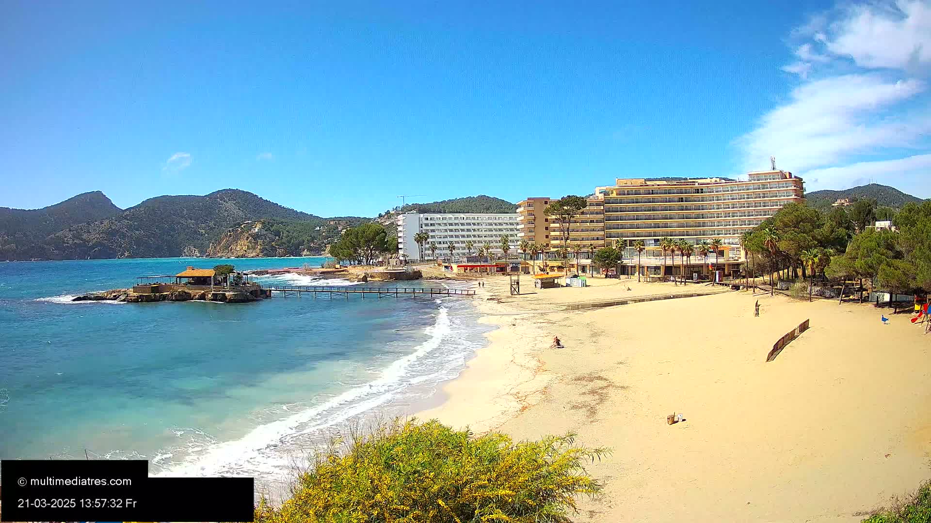A sandy beach with a hotel and a small building on a rock in the water, under a clear blue sky with a few clouds.