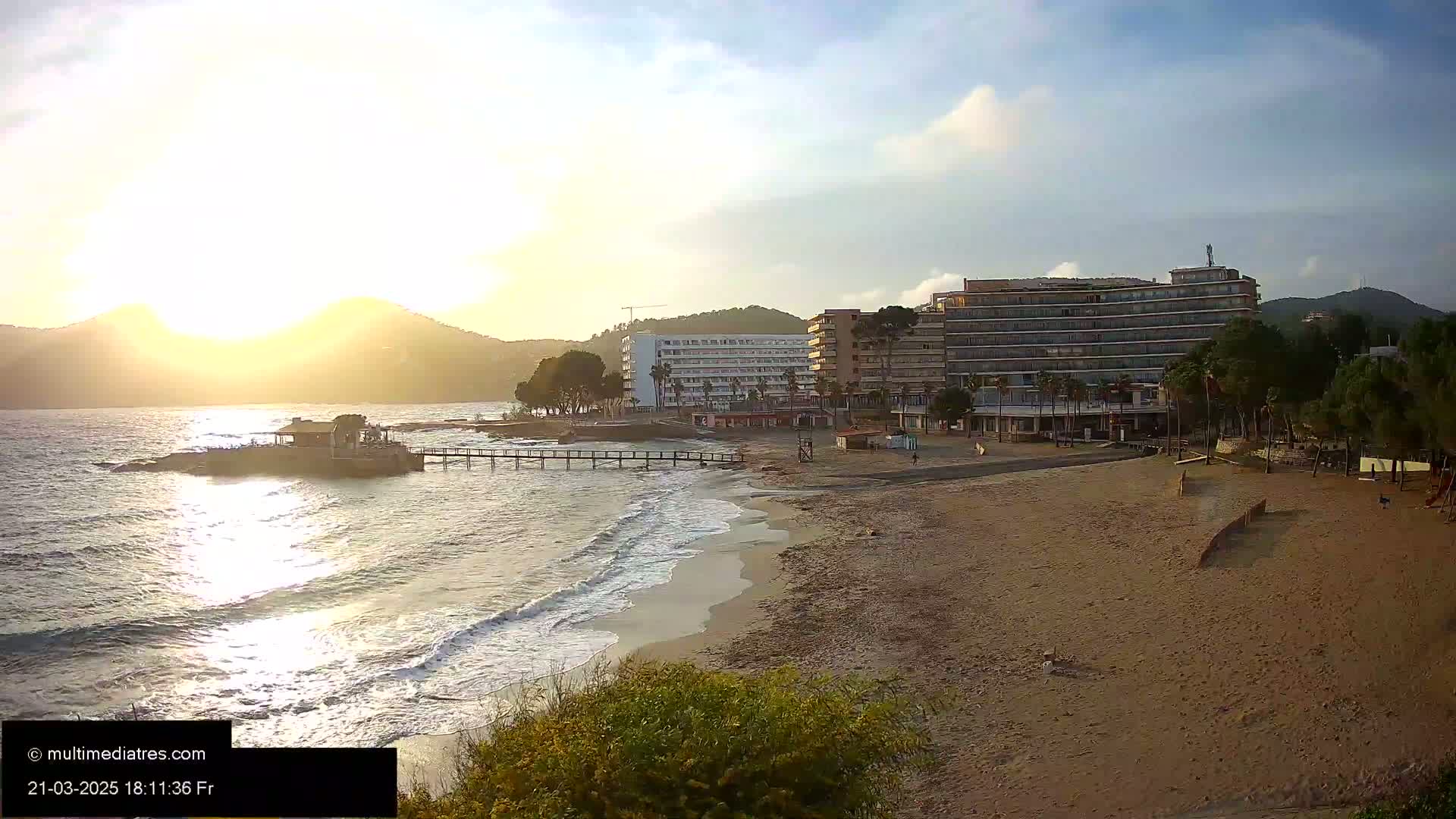 A sandy beach with gentle waves laps at the shore under a partly cloudy sunset sky, with several hotels visible in the background.