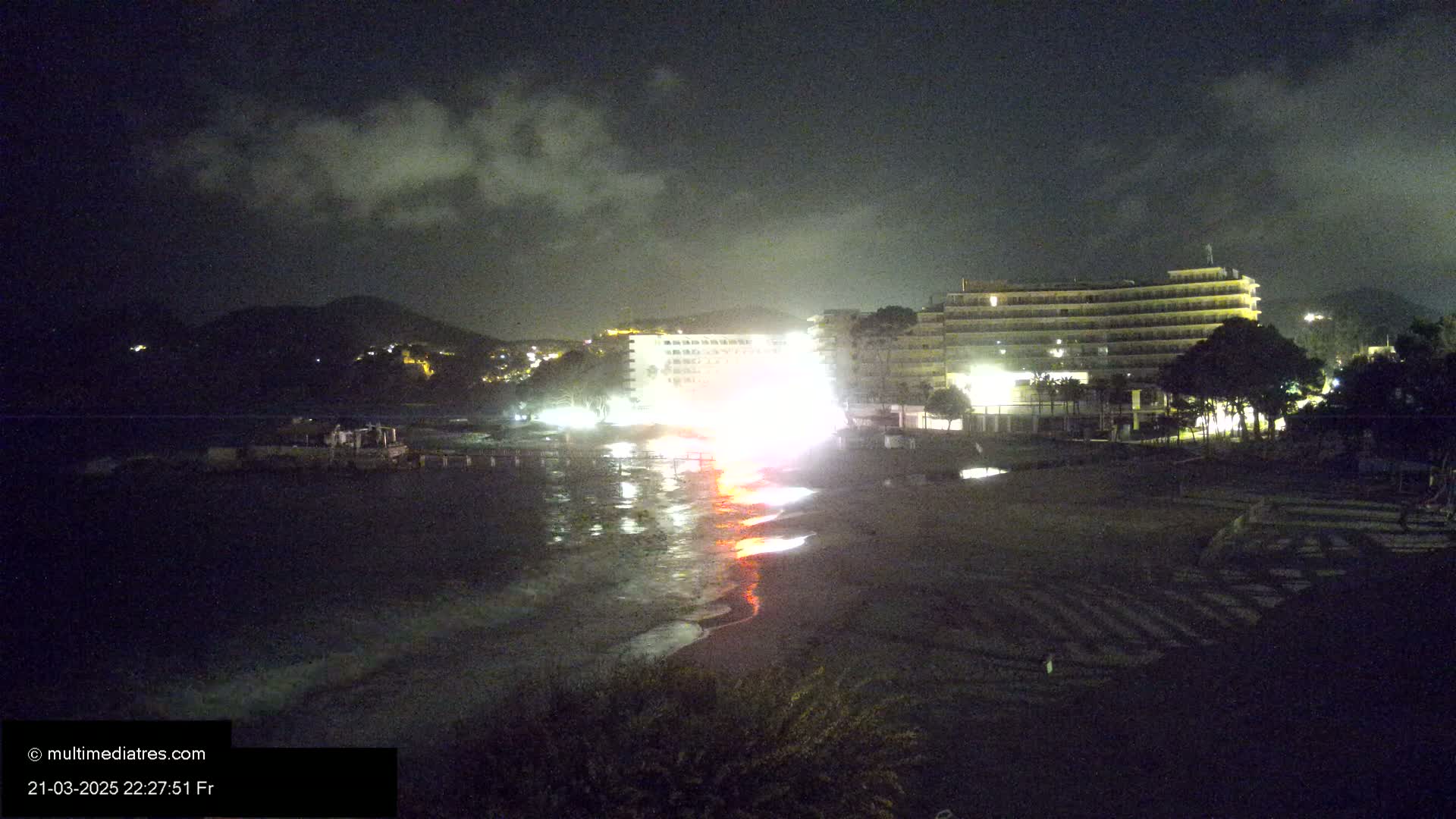 A nighttime view of a beach with a multi-story hotel and a hill in the background under a partly cloudy sky.