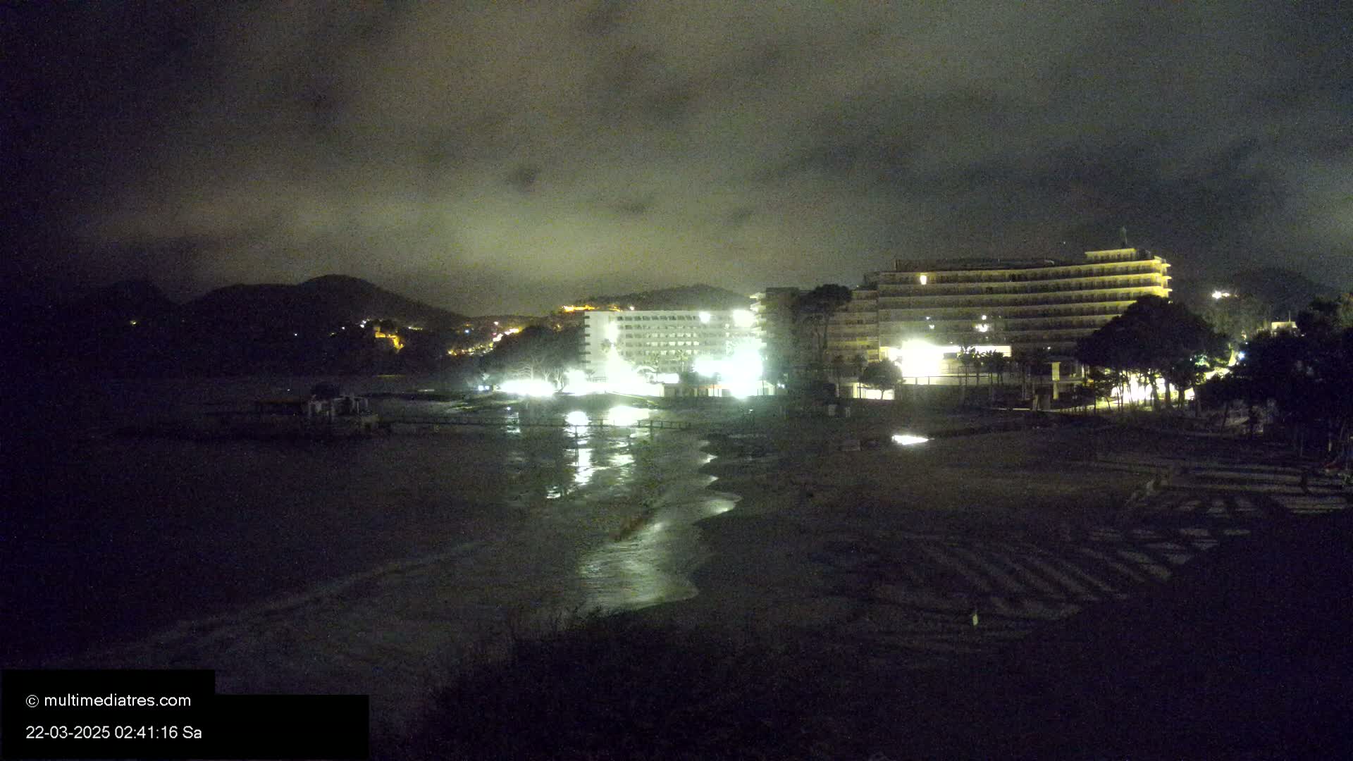 At night, under a cloudy sky, illuminated hotels stand near a dark beach with a calm, reflective body of water.