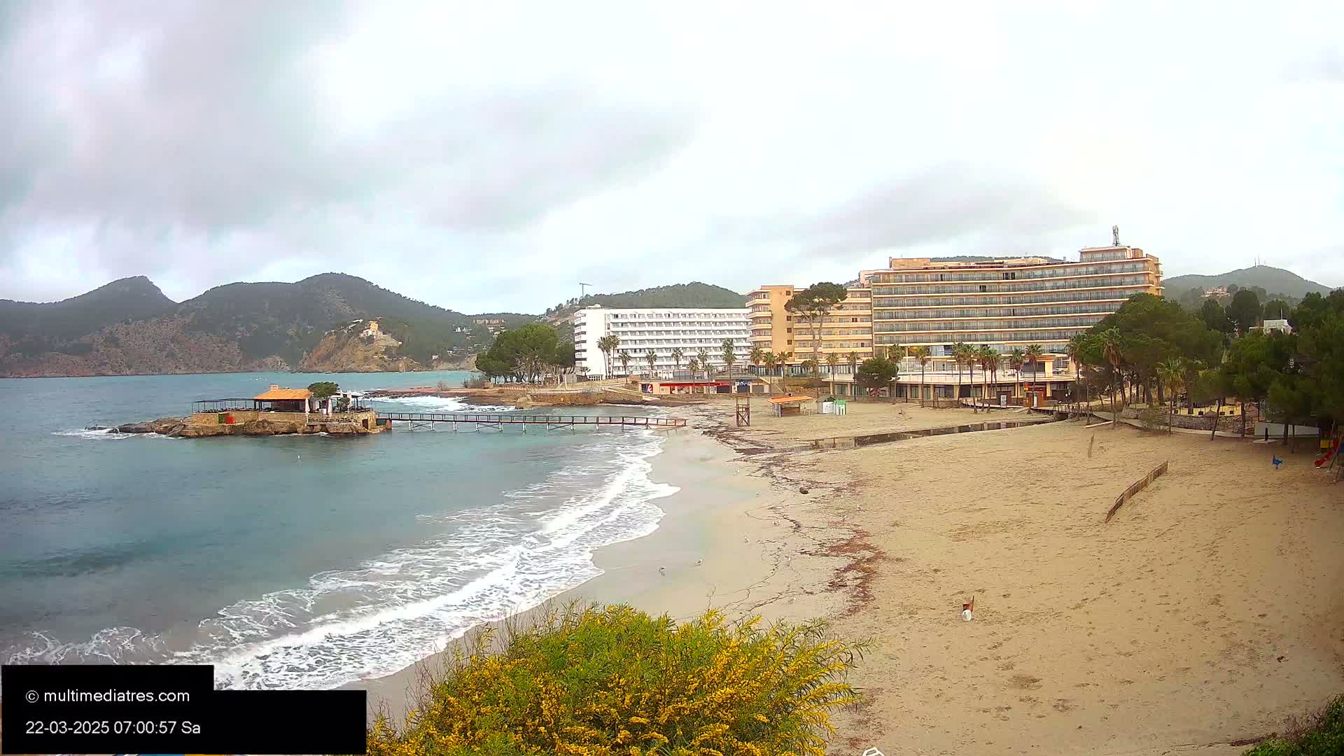A sandy beach with gentle waves laps against the shore,  under a cloudy sky,  with hotels and vegetation visible in the background.