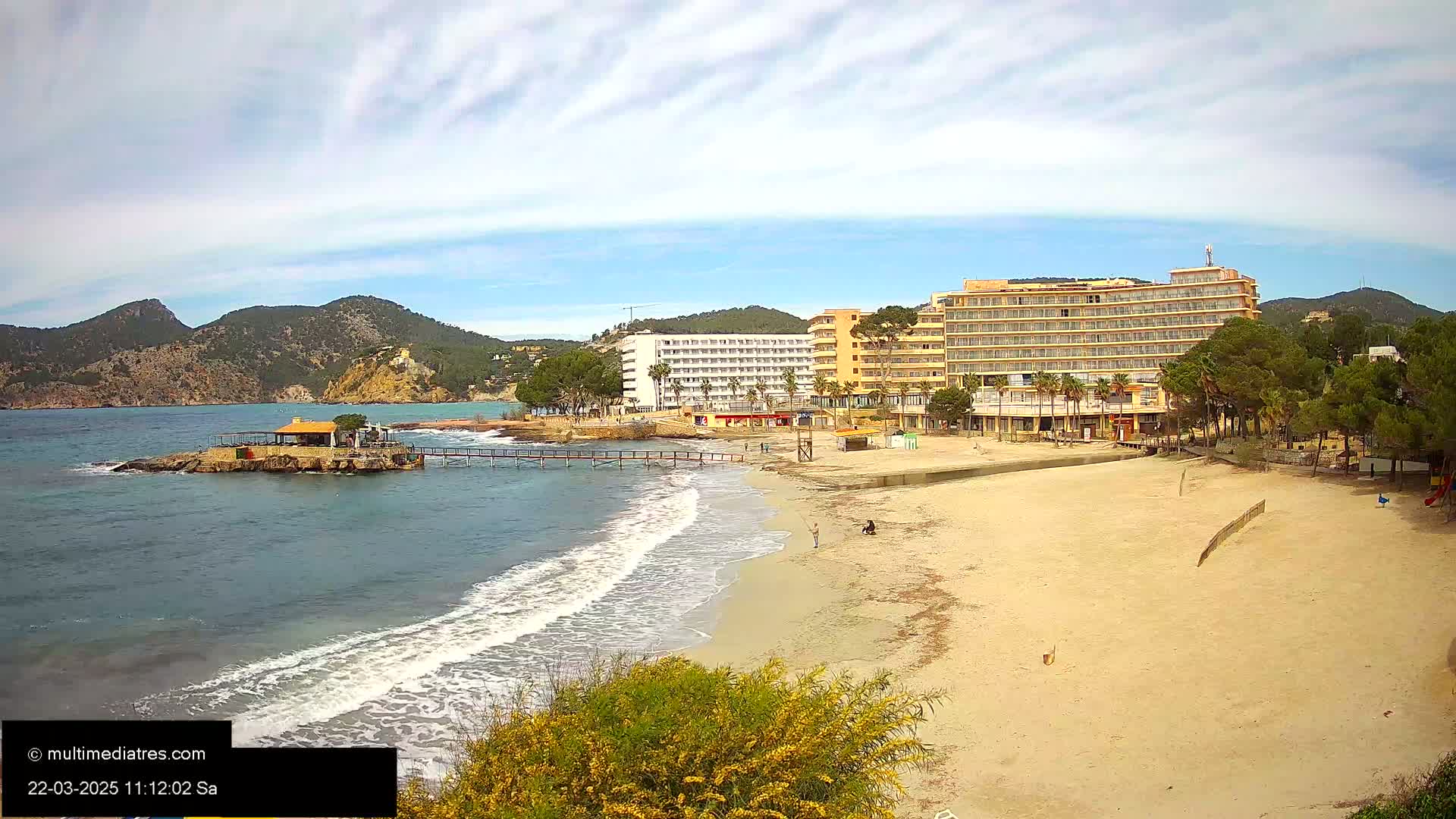 A sandy beach with gentle waves laps against the shore near two large hotels and a small island with a structure, all under a partly cloudy sky.