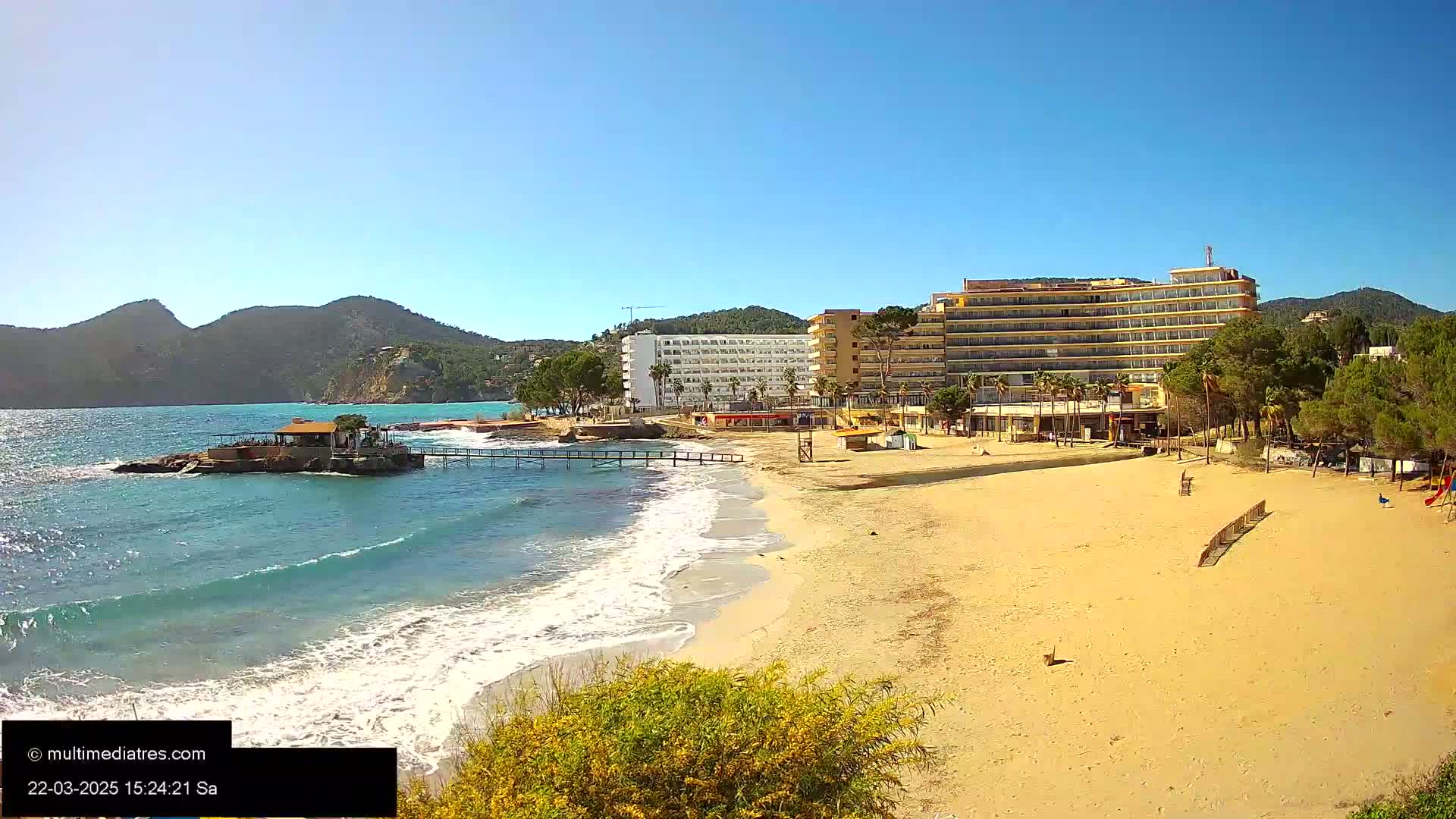 A sandy beach with gentle waves laps against the shore, near a beachfront hotel and a small restaurant on a rocky outcrop under a clear blue sky.