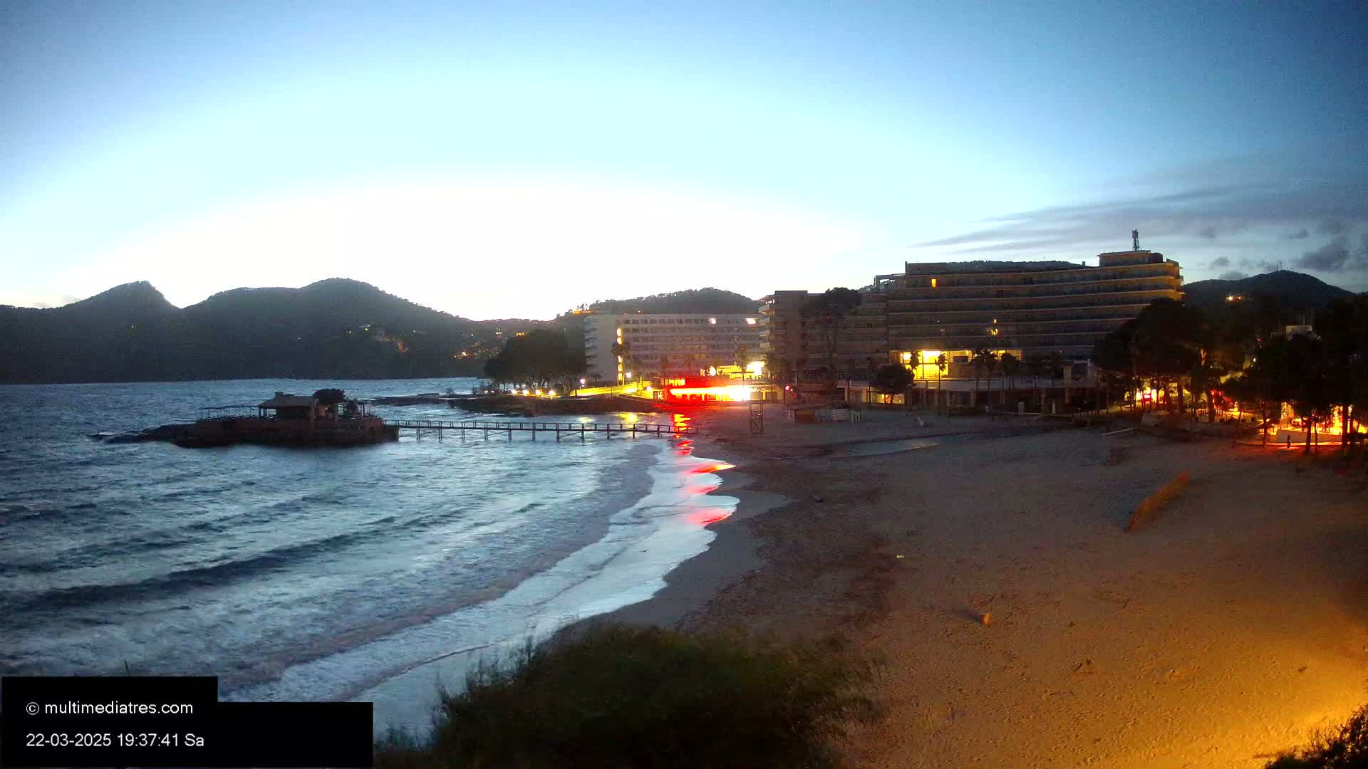 A sandy beach at dusk, with gently lapping waves, is bordered by a row of buildings illuminated against a backdrop of dark mountains under a twilight sky.