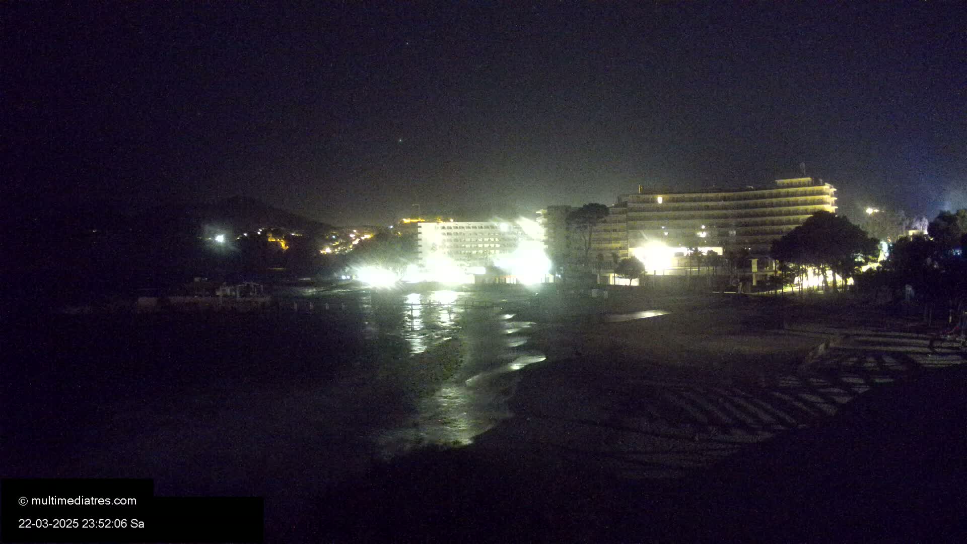 Lit-up hotels and buildings stand on a dark beach at night, with gentle waves lapping at the shore under a clear sky.