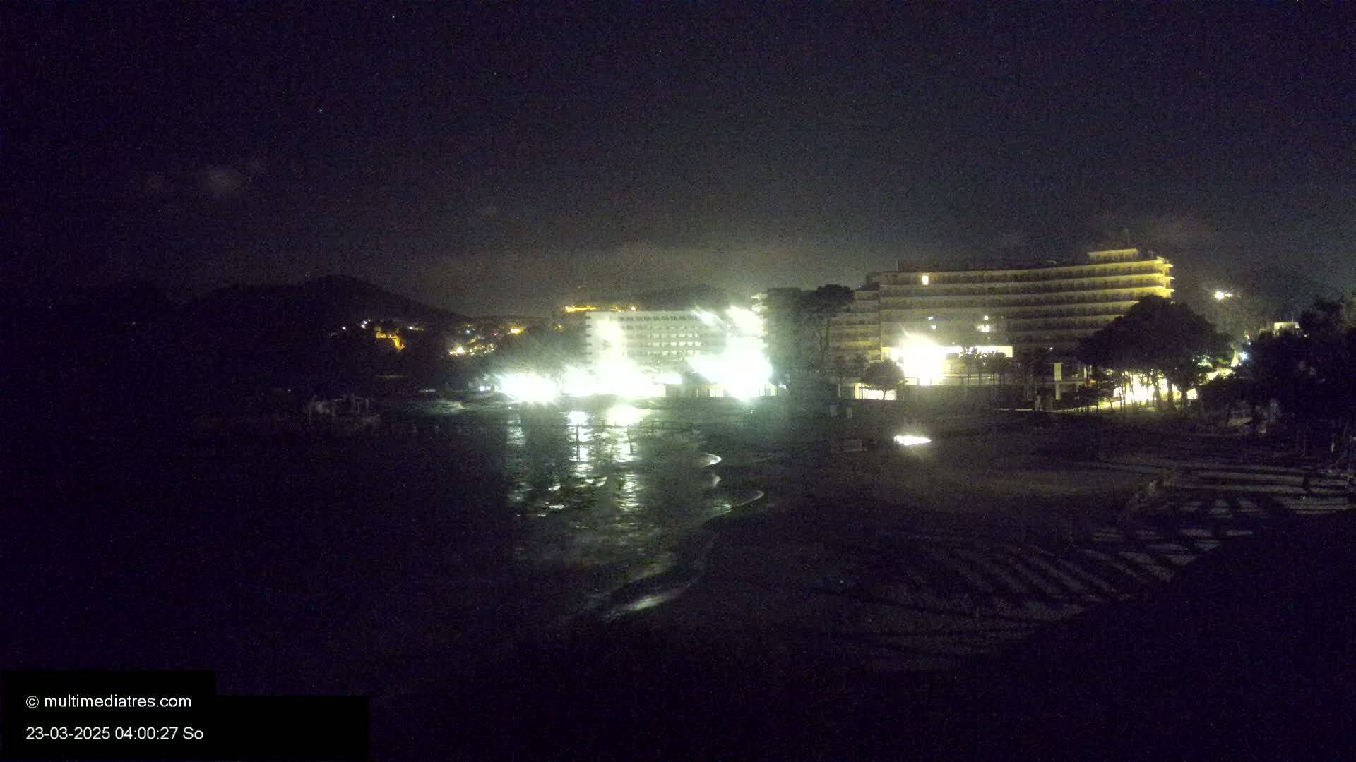 At night, a dark beach with gently lapping waves is adjacent to brightly lit hotels and a hill in the distance under a mostly clear sky.