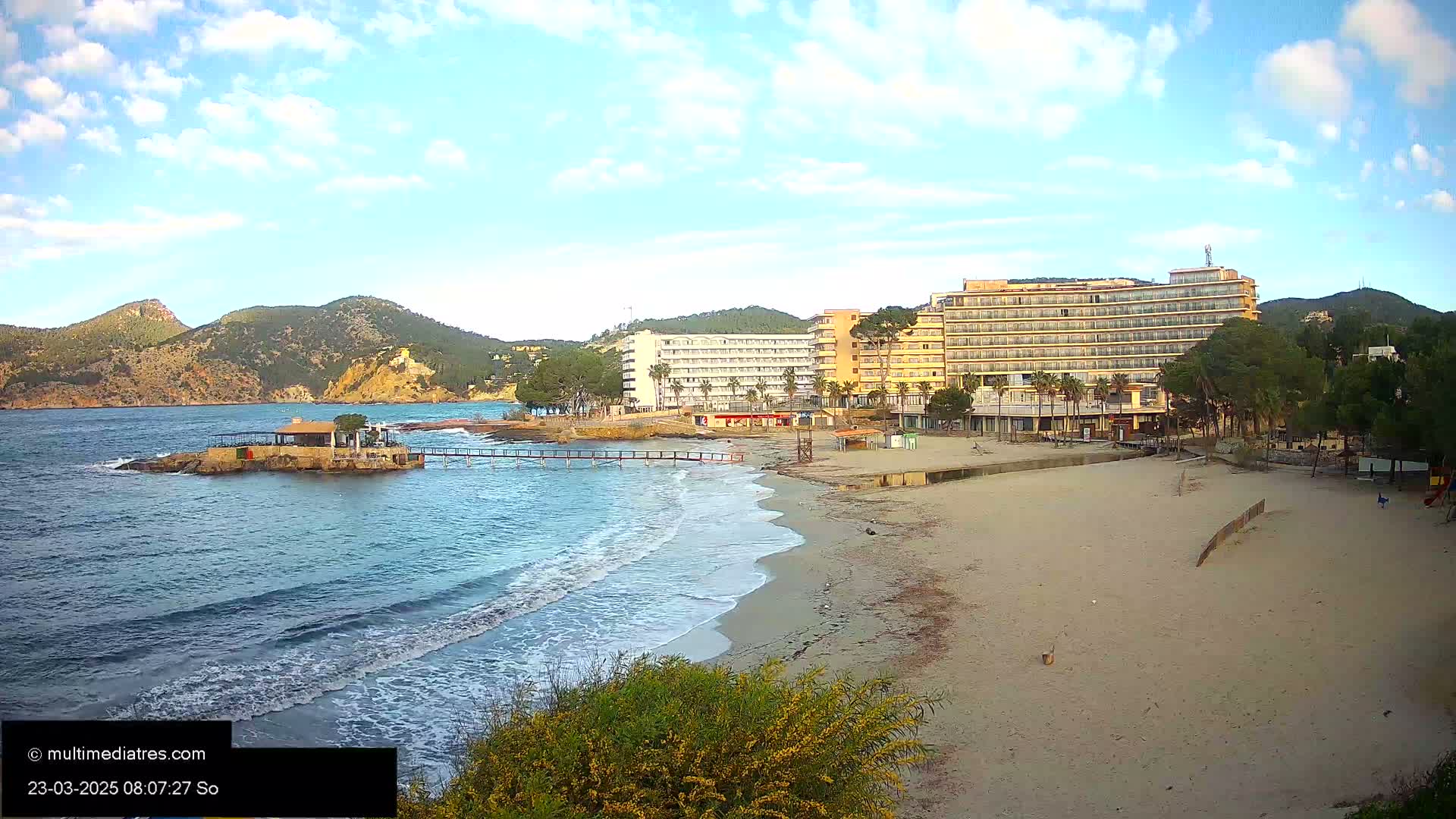 A sandy beach with small waves laps against the shore, near a couple of hotels and a small island with a building under a partly cloudy sky.