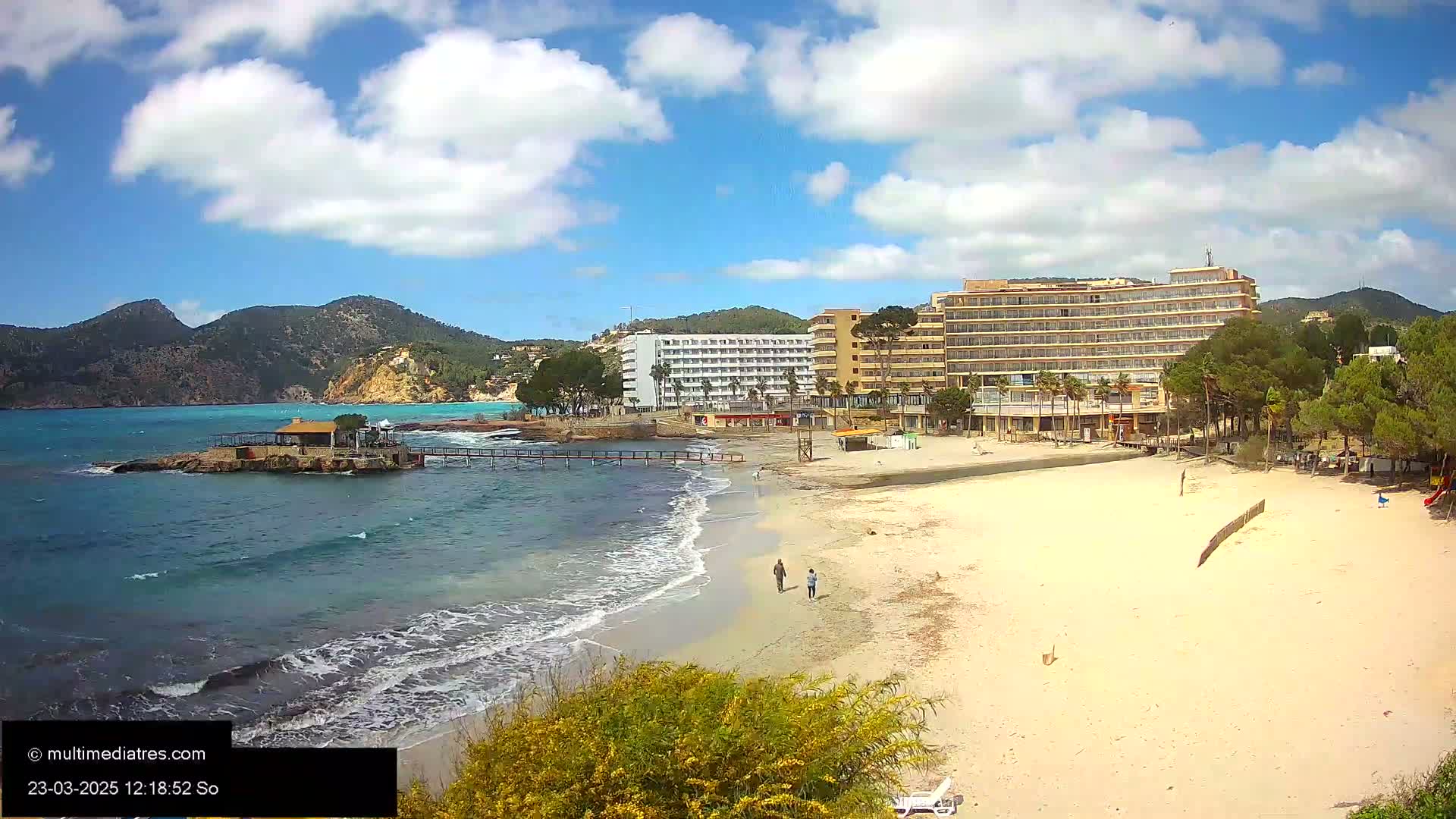 A sandy beach with a few people walking near a hotel and some buildings, backed by hills, under a partly cloudy blue sky.