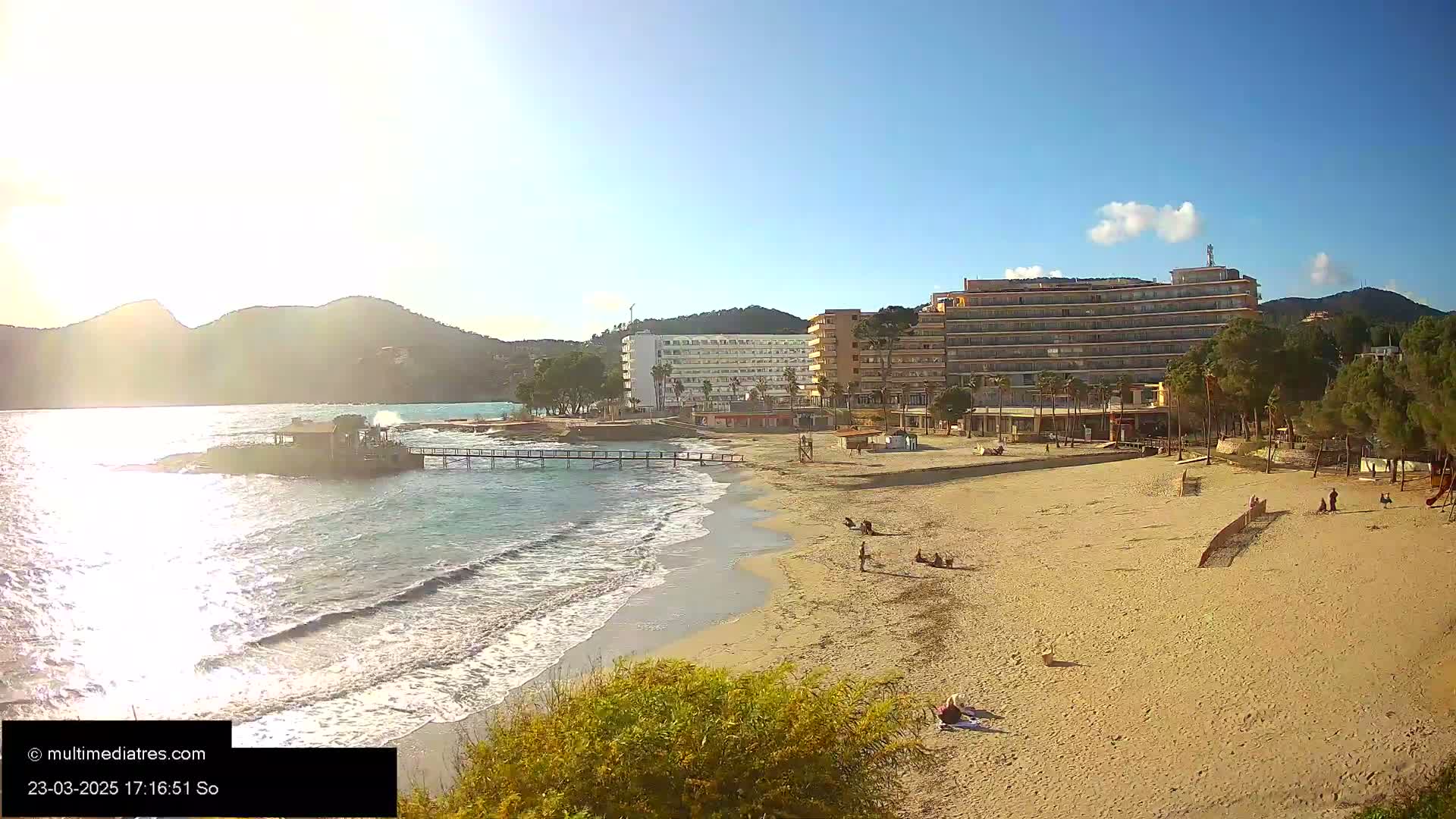 A sandy beach with gentle waves laps the shore near several hotels and a pier, under a mostly sunny sky with a few clouds.