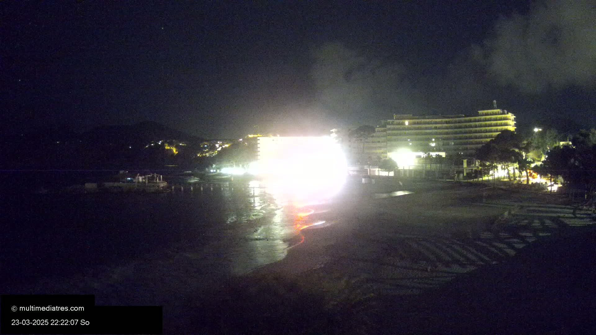 A bright light illuminates a beach at night near a large hotel, with dark clouds in the sky.