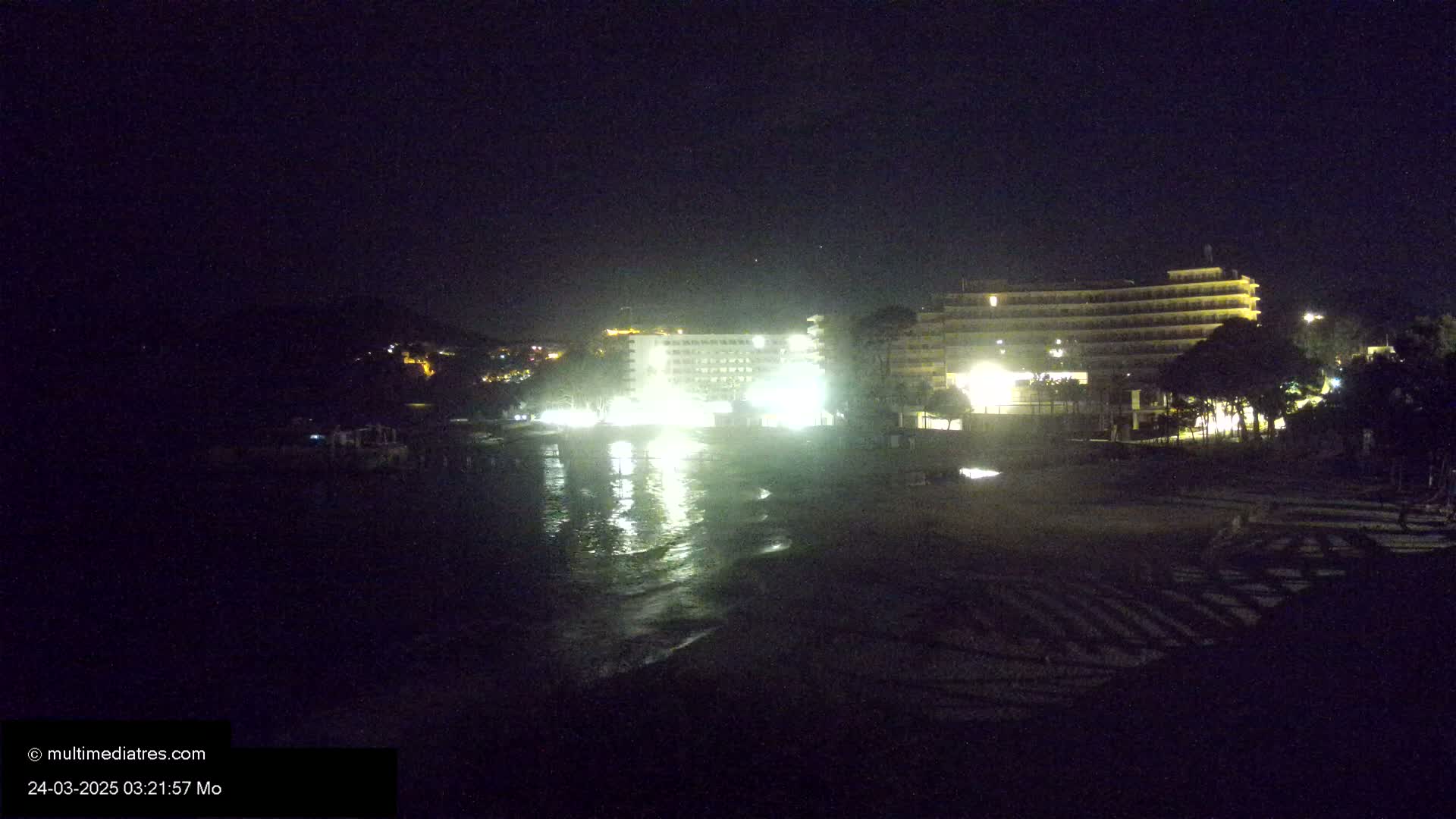 At night, a dark beach with gently lapping waves reflects the lights of nearby hotels and buildings on a calm sea.