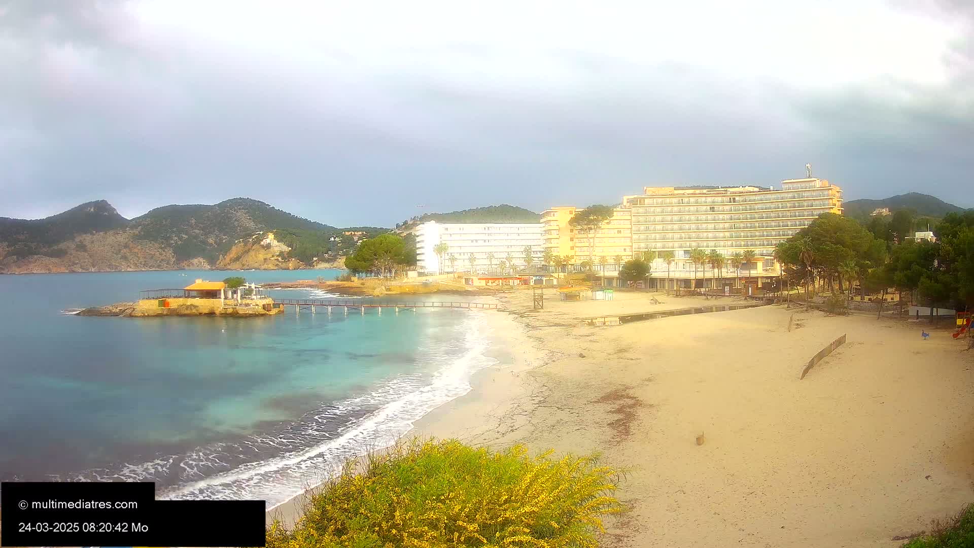A sandy beach with calm, turquoise water laps at its edge, in front of several hotels and a small island with a pavilion, all under a mostly cloudy sky.
