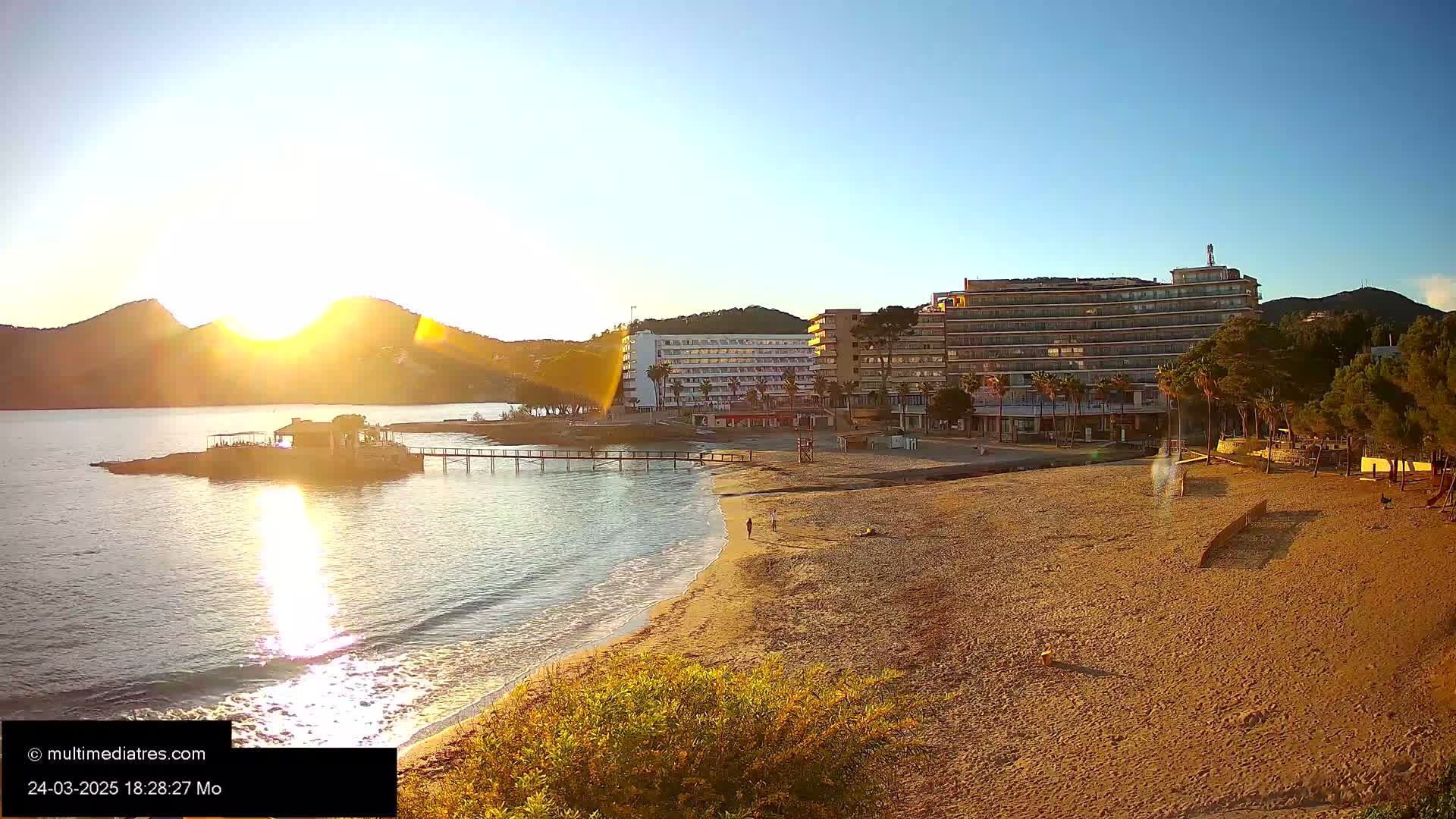 A sandy beach and calm ocean are seen at sunset, with a hotel and pier in the background.