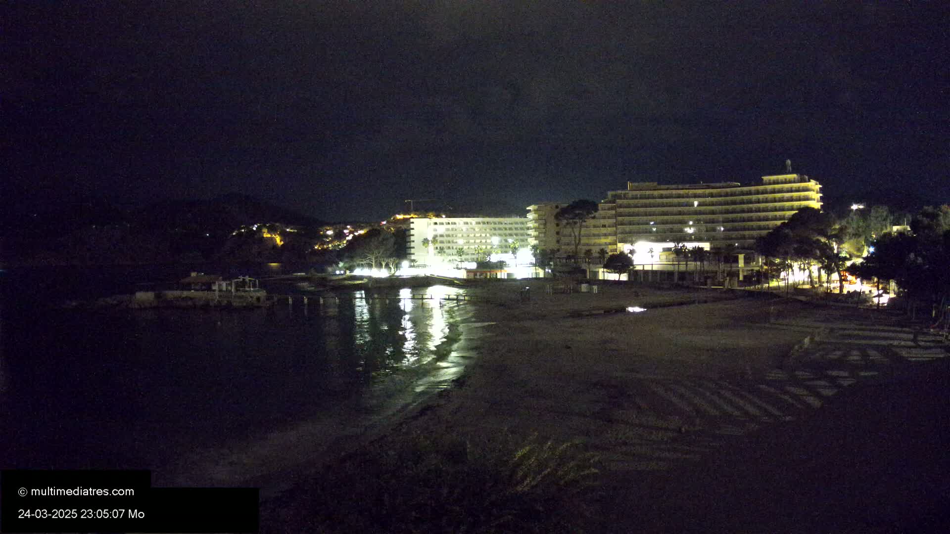 A nighttime view of a beach with two illuminated hotels and a calm, reflective body of water.