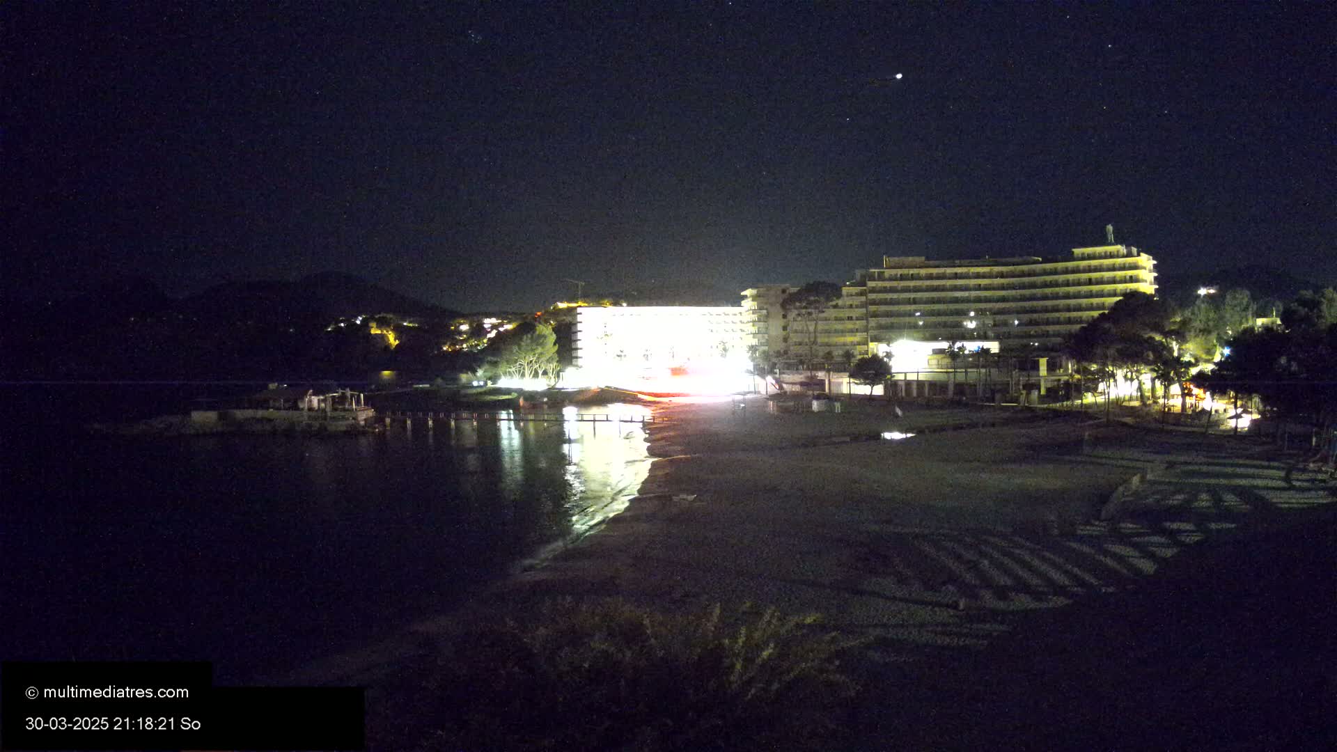 A brightly lit beachfront hotel is visible at night under a clear, starry sky next to a dark body of water and a sandy beach.