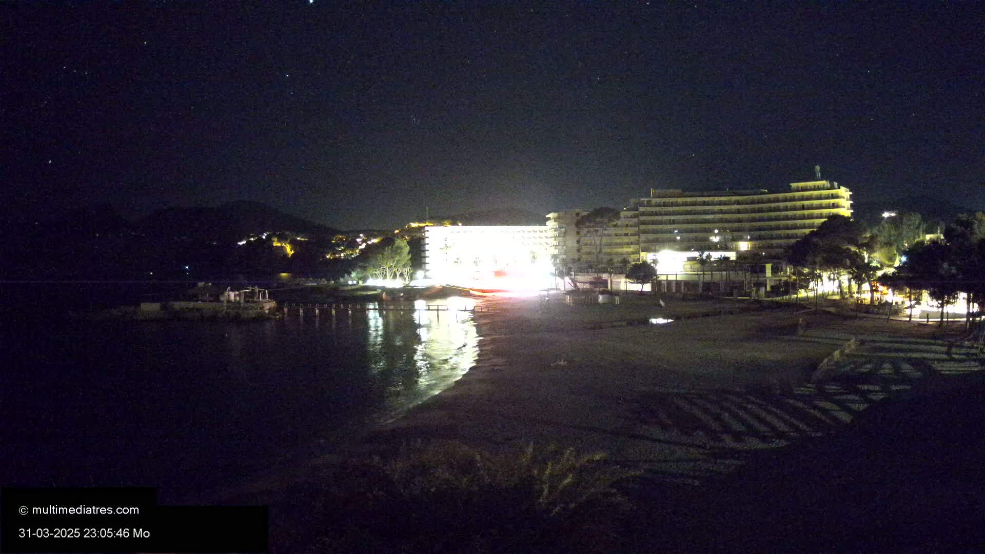 A brightly lit hotel complex sits on a dark beach at night under a starry sky, next to calm, reflective water.
