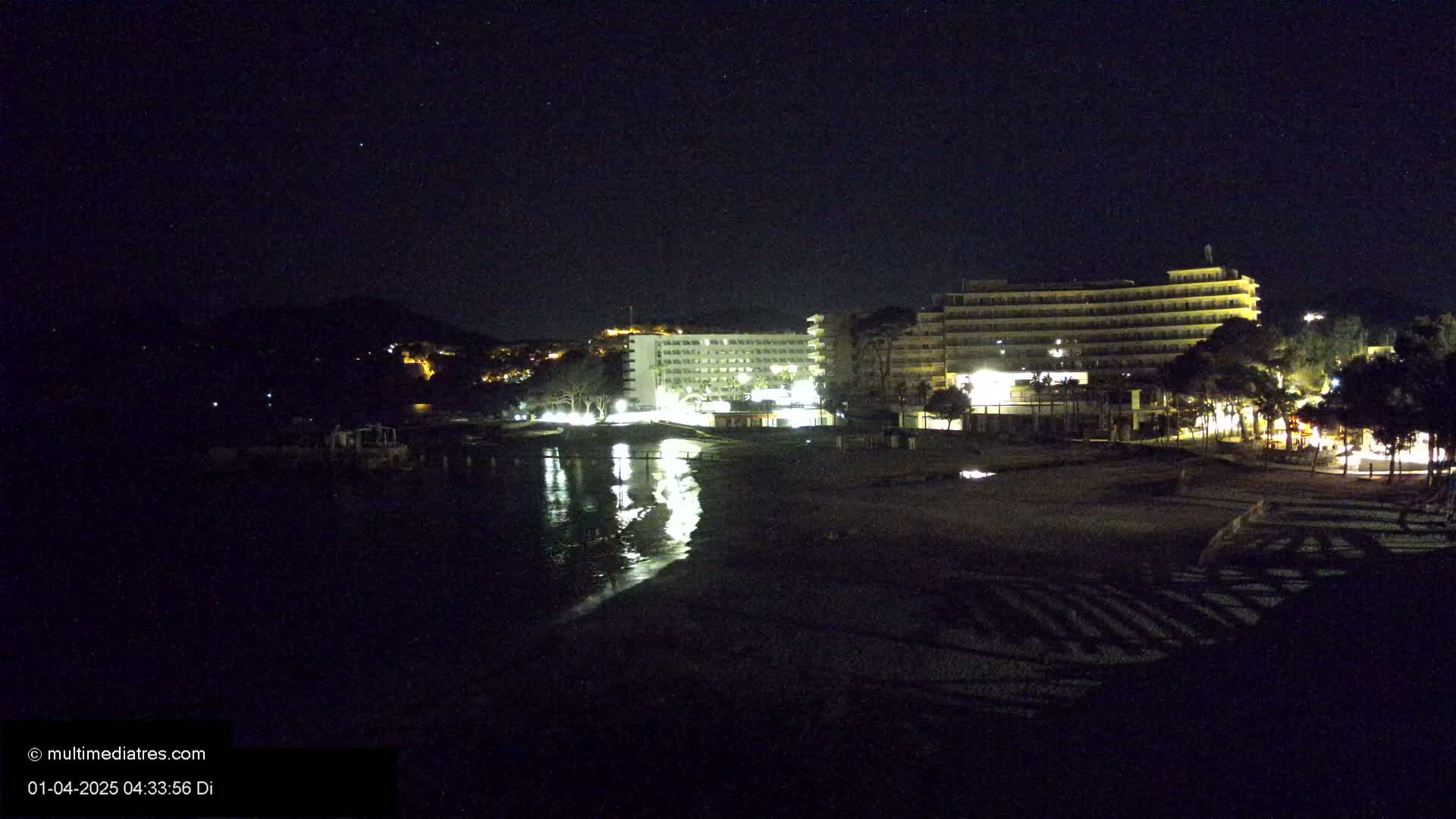 At night, two illuminated hotels are situated near a dark beach and calm water, under a clear, starry sky.
