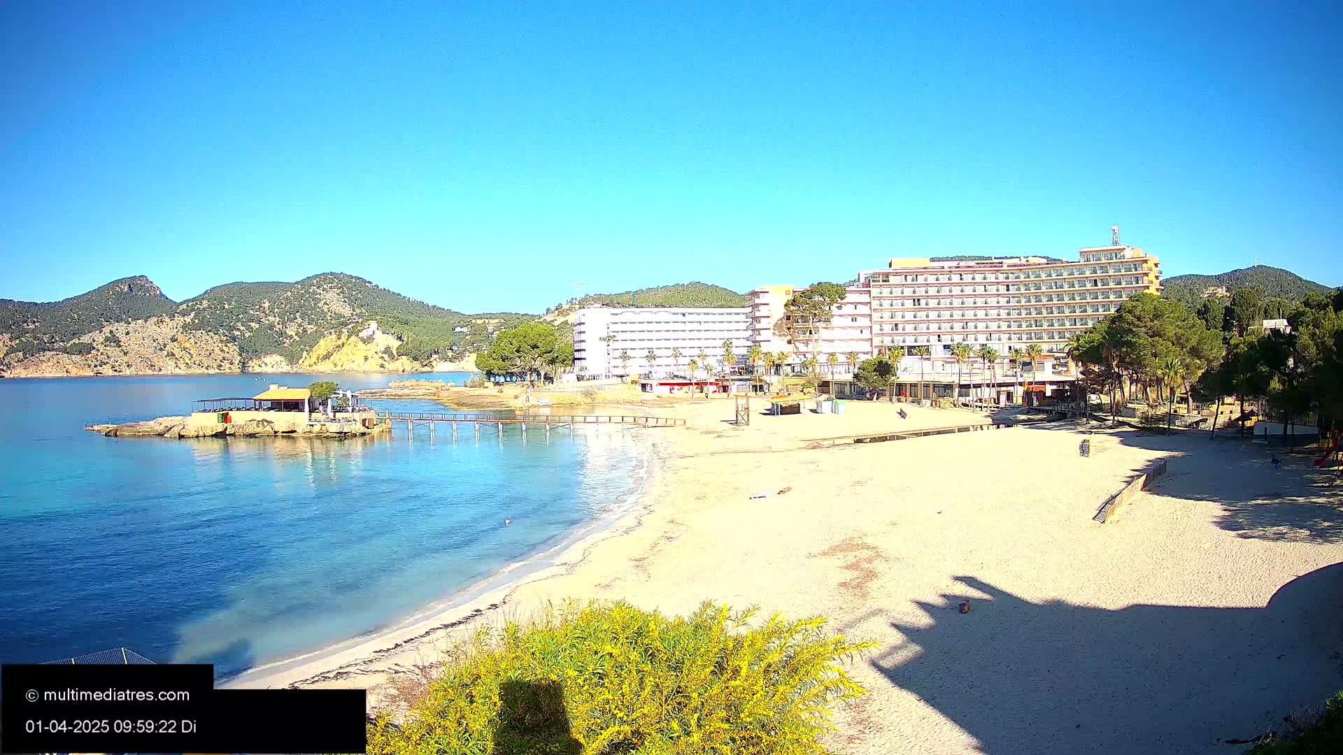 A sandy beach with clear blue water, a beachfront hotel, and a small restaurant on a rock outcropping, all under a bright sunny sky.
