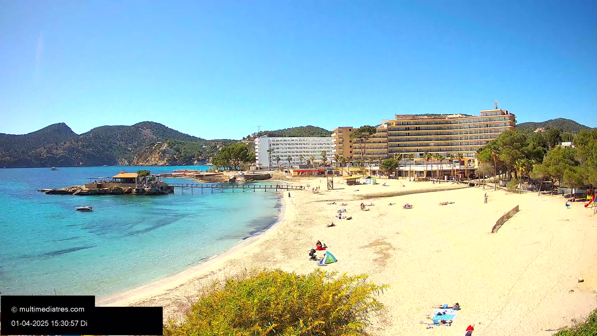 A sandy beach with turquoise water, a few sunbathers, hotels, and a small pier extends towards a calm, clear ocean under a bright sunny sky.