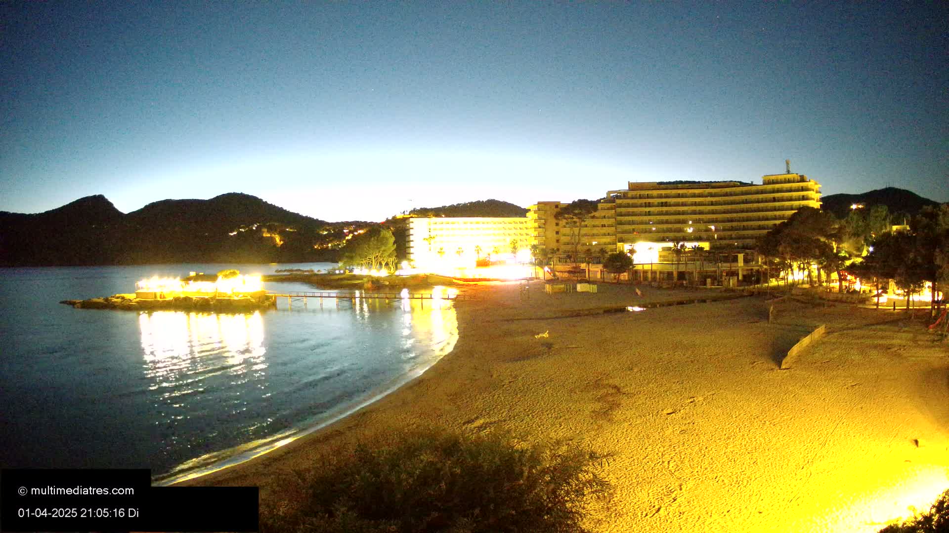 A brightly lit beach at night, with a large hotel and calm water reflecting the lights, under a dark blue twilight sky.
