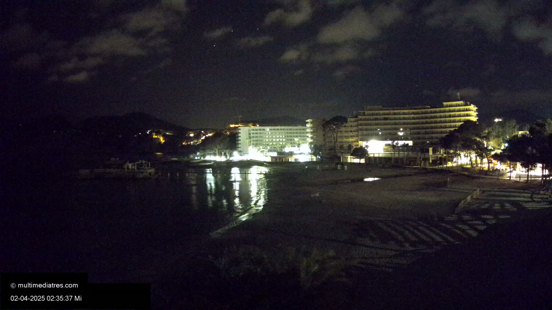 At night, under a partly cloudy sky, illuminated hotels and buildings are situated near a dark beach and calm body of water, with a hill visible in the background.