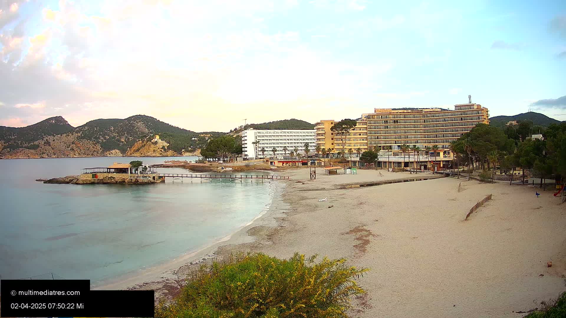 A sandy beach with calm, light-blue water stretches out in front of several hotels and a small building on a rocky outcropping, all under a partly cloudy sky.