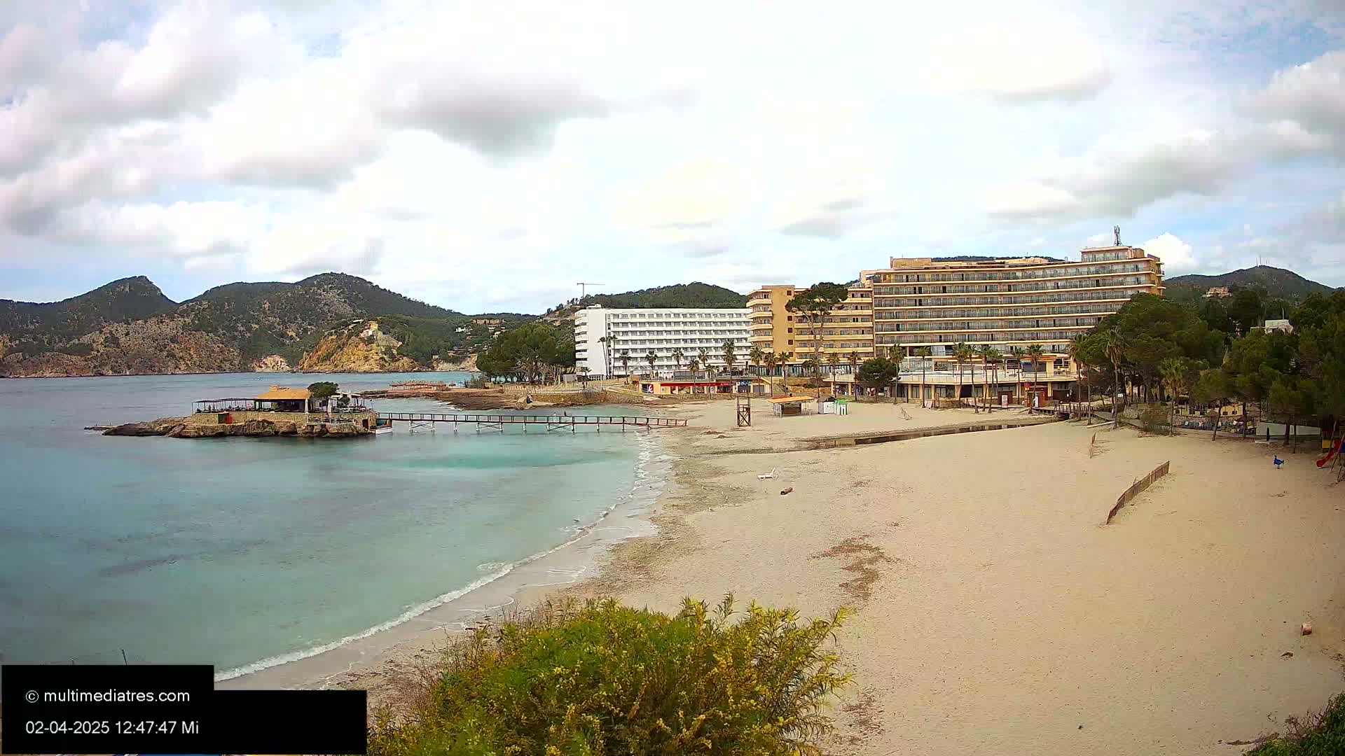 A sandy beach with calm, light-blue water stretches out in front of several hotels and a small restaurant situated on a rocky outcrop, all under a partly cloudy sky.