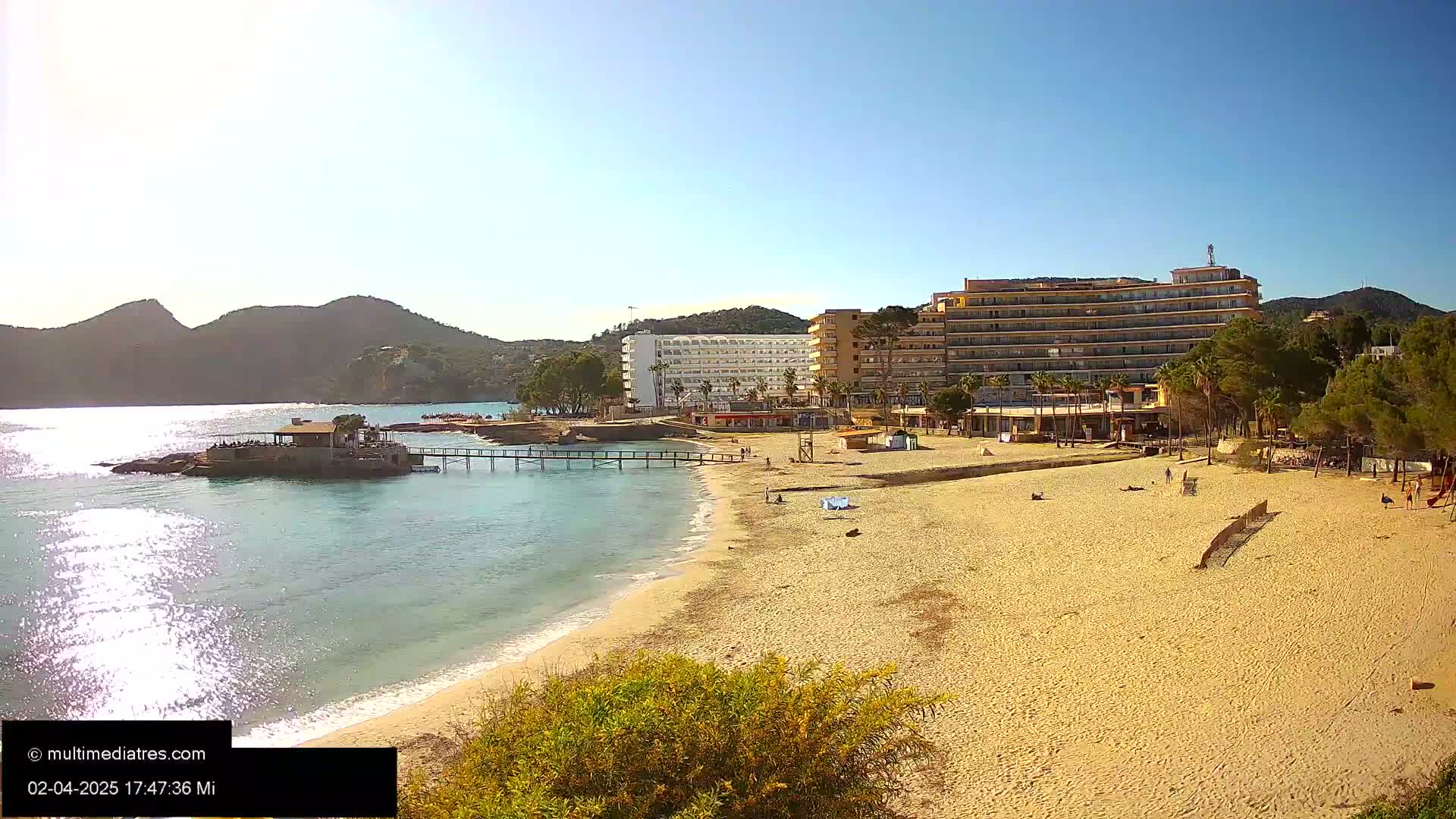 A sandy beach with a few people on it stretches toward hotels and a pier, all backed by hills under a sunny sky.