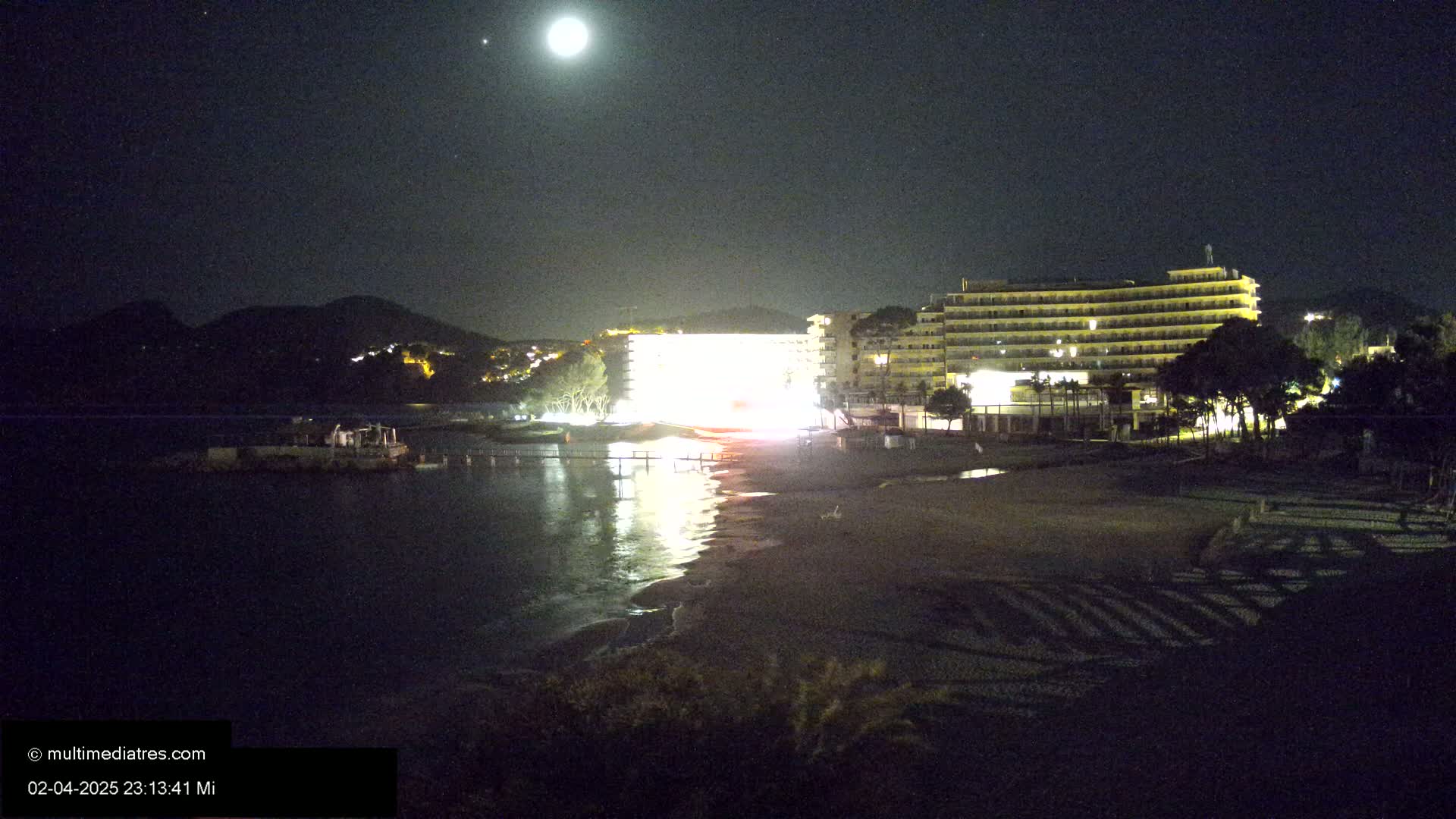 A brightly lit hotel sits on a beach at night under a full moon, with calm water reflecting the lights.