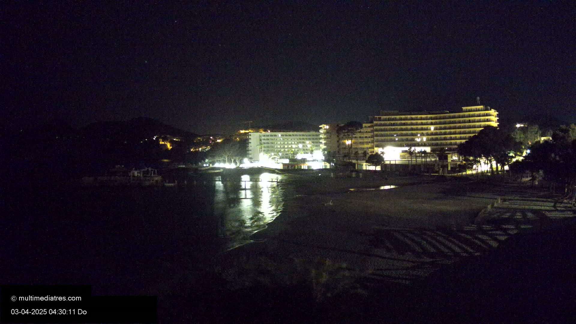 A nighttime view of a beach with two illuminated hotels and calm water reflecting the lights.