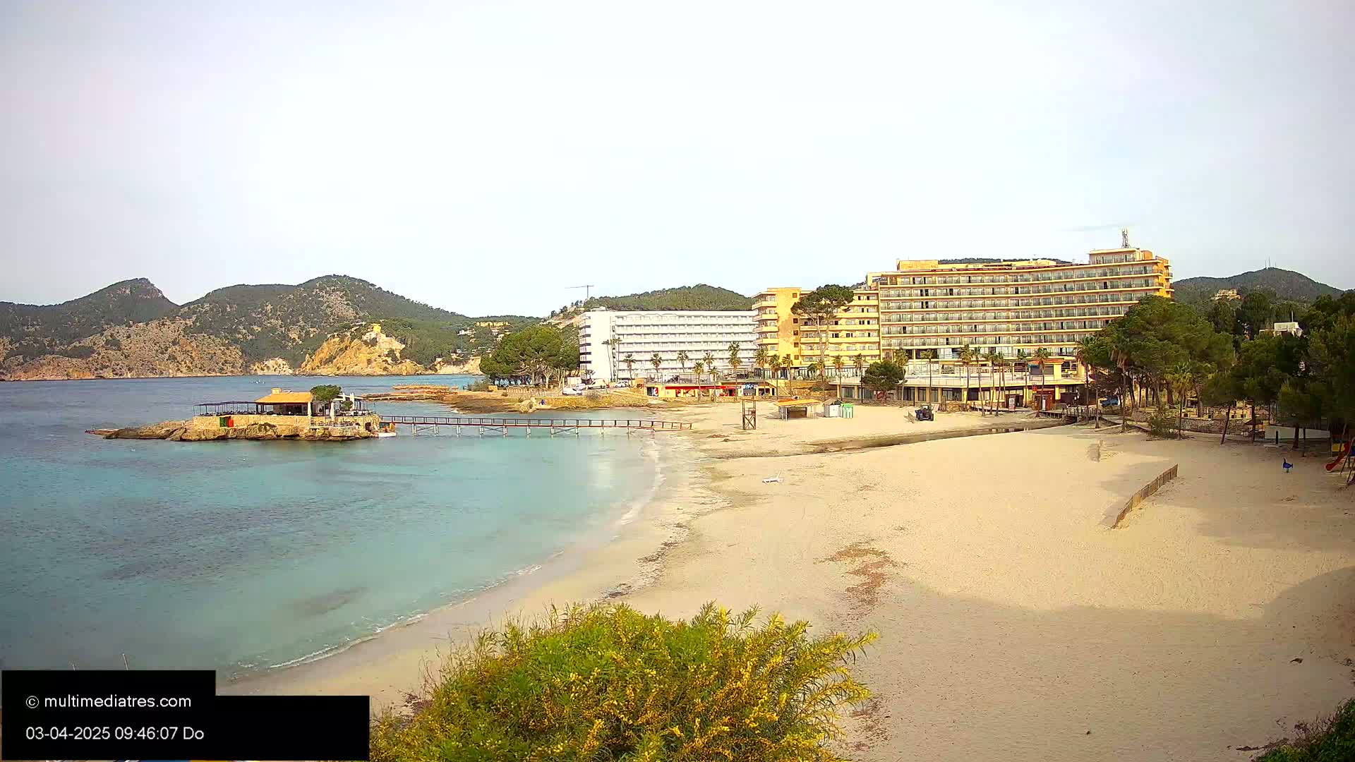 A sandy beach with calm, turquoise water stretches toward two large hotels and a small restaurant on a rocky outcropping, all backed by low, green hills under a bright, clear sky.