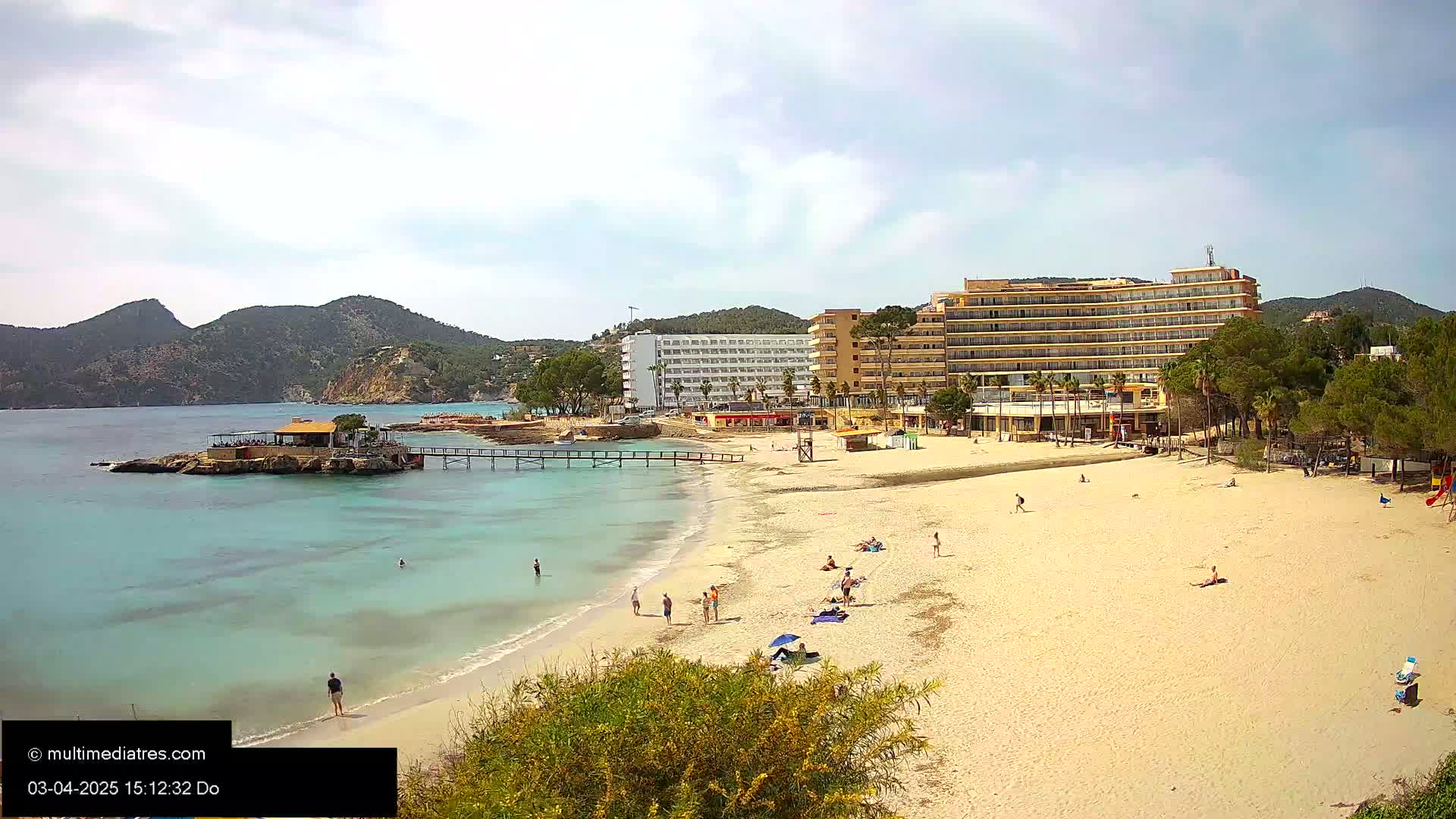 A sandy beach with a few people on it, a small pier extending into clear turquoise water, and several hotels and buildings behind it under partly cloudy skies.