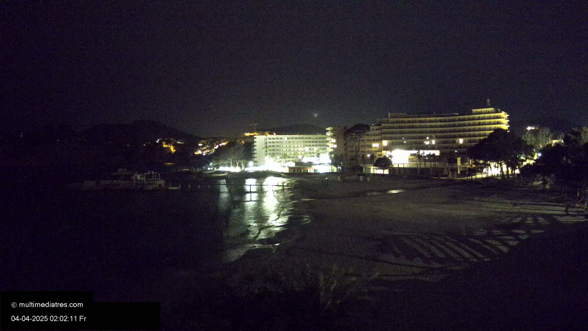 A nighttime view of a beach with two illuminated hotels, a calm body of water, and dark vegetation.