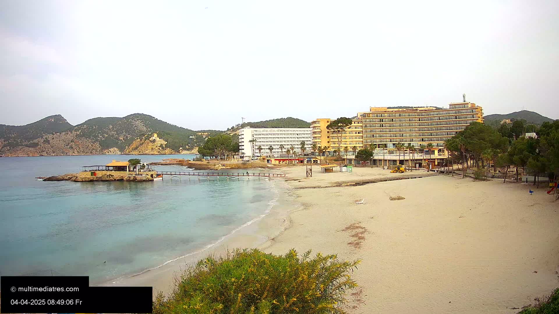 A sandy beach with calm, turquoise water stretches out before two large hotels and a small building on a rocky outcropping under an overcast sky.