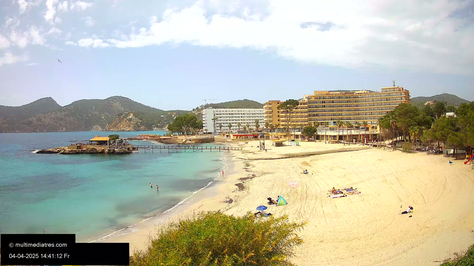 A sandy beach with turquoise water, several hotels, and people sunbathing under a partly cloudy sky.