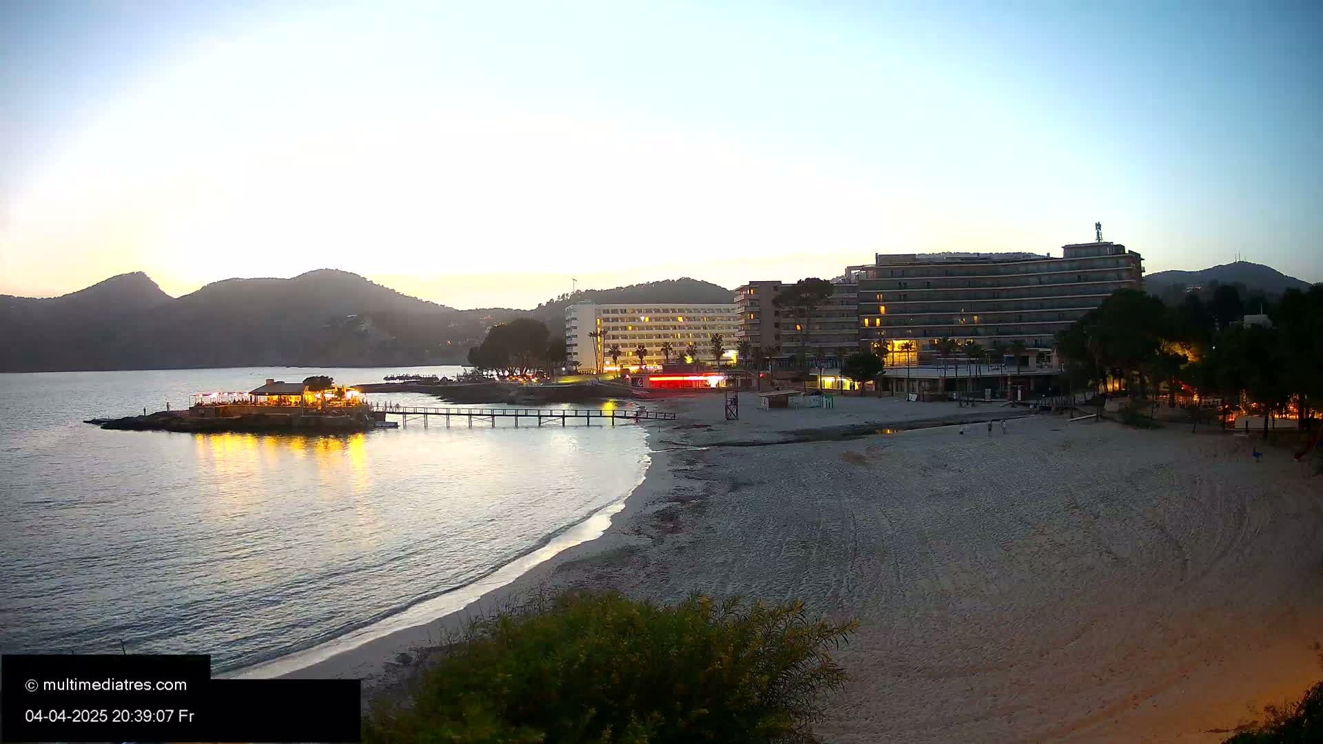 A sandy beach at dusk, with a lit-up beachfront restaurant on a small island, a pier, and several hotels behind it, all under a clear twilight sky.